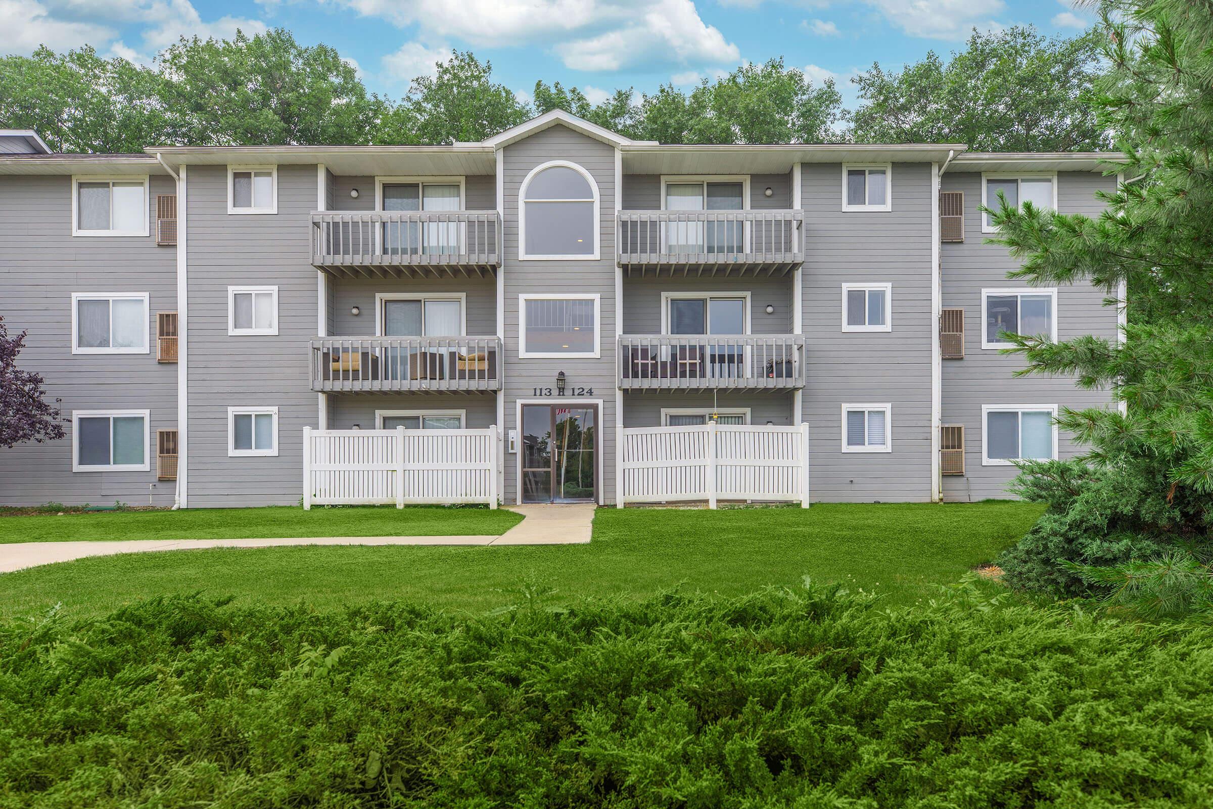 Exterior view of a multi-story apartment building with gray siding. The building features balconies, large windows, and a white picket fence in front. Lush green grass and shrubs surround the property, under a partly cloudy sky.