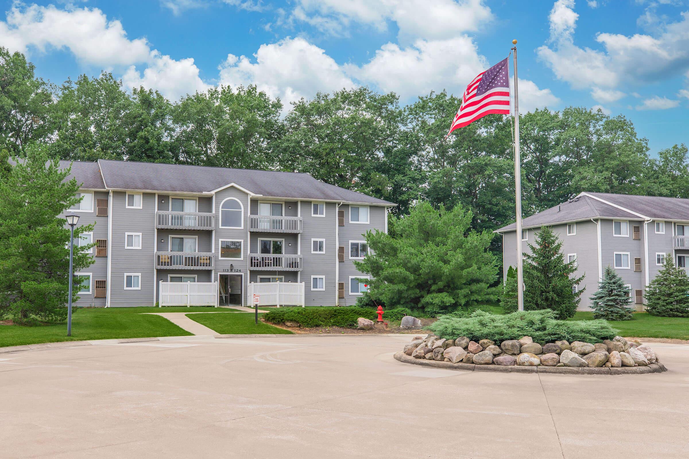 A residential apartment complex featuring two buildings with balconies, surrounded by lush greenery. A circular driveway with a stone feature in the center and an American flag fluttering in front. The sky is partly cloudy, adding to the tranquil atmosphere.