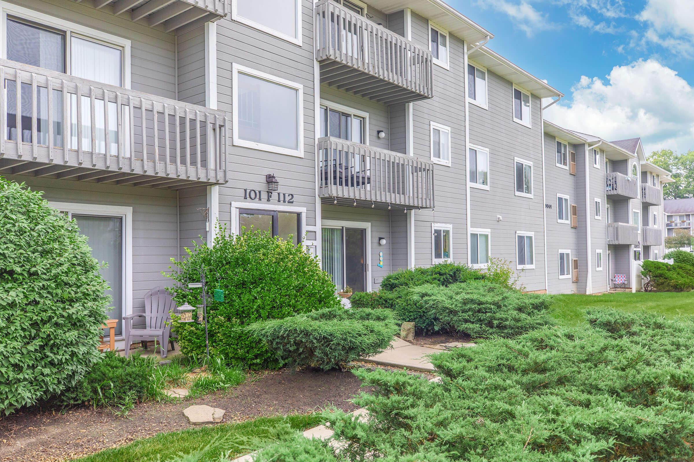 A multi-unit apartment building with gray siding and multiple balconies. The foreground features well-maintained shrubbery and a stone path leading to the entrance. The sky is partly cloudy, adding a bright atmosphere to the scene.