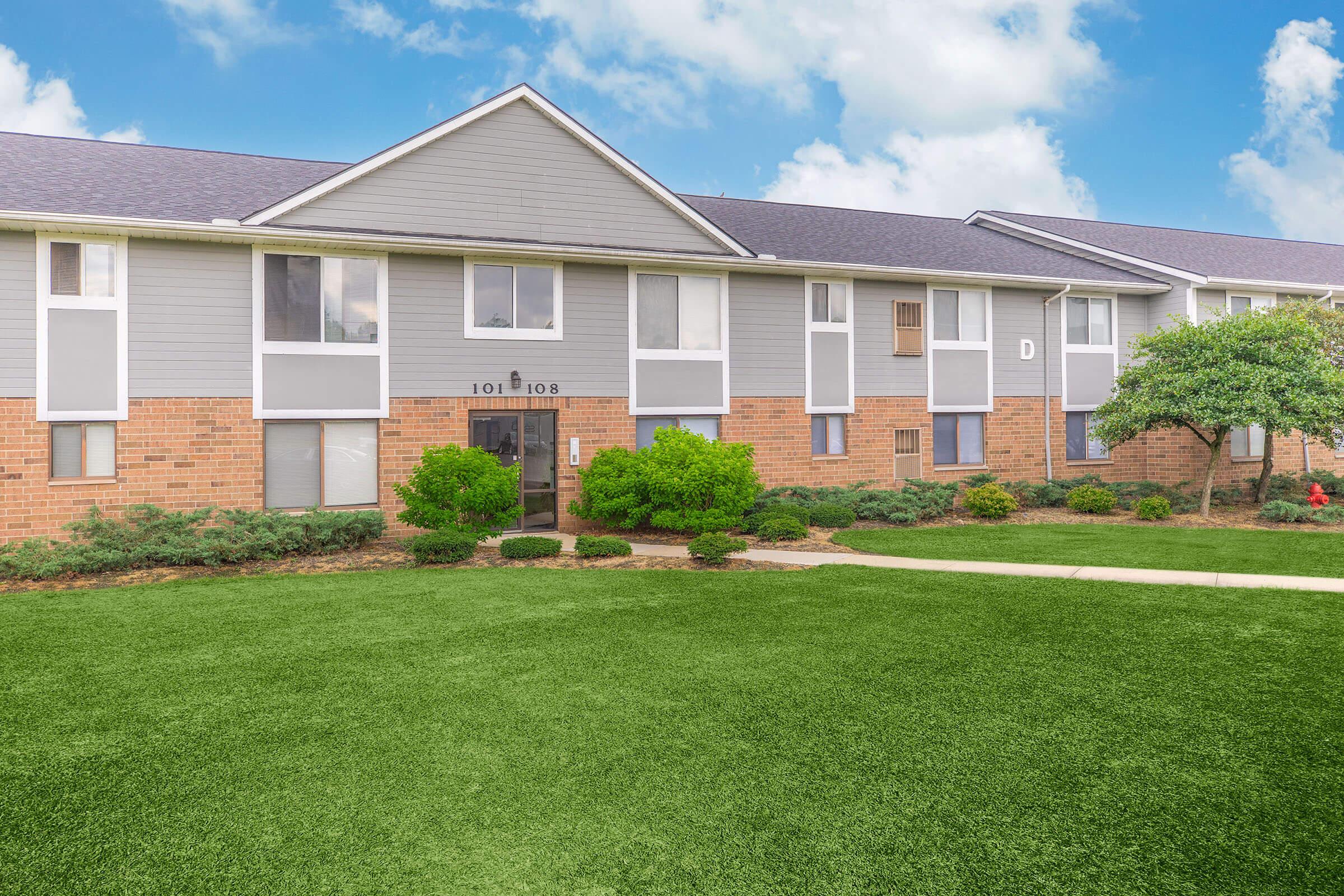 A two-story residential building with a brick facade and light gray siding, surrounded by neatly manicured green grass and bushes. The building features several windows and an entrance marked with the numbers 101 and 108, under a blue sky with fluffy clouds.