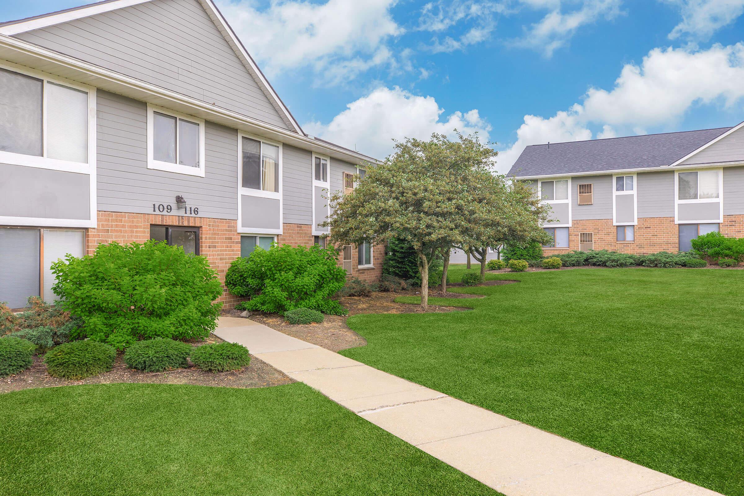 Exterior view of a residential building complex featuring two buildings on either side of a manicured green lawn. A paved walkway leads through neatly trimmed shrubs and small trees, under a partly cloudy sky, creating an inviting atmosphere. The buildings have a mix of gray and brick siding.