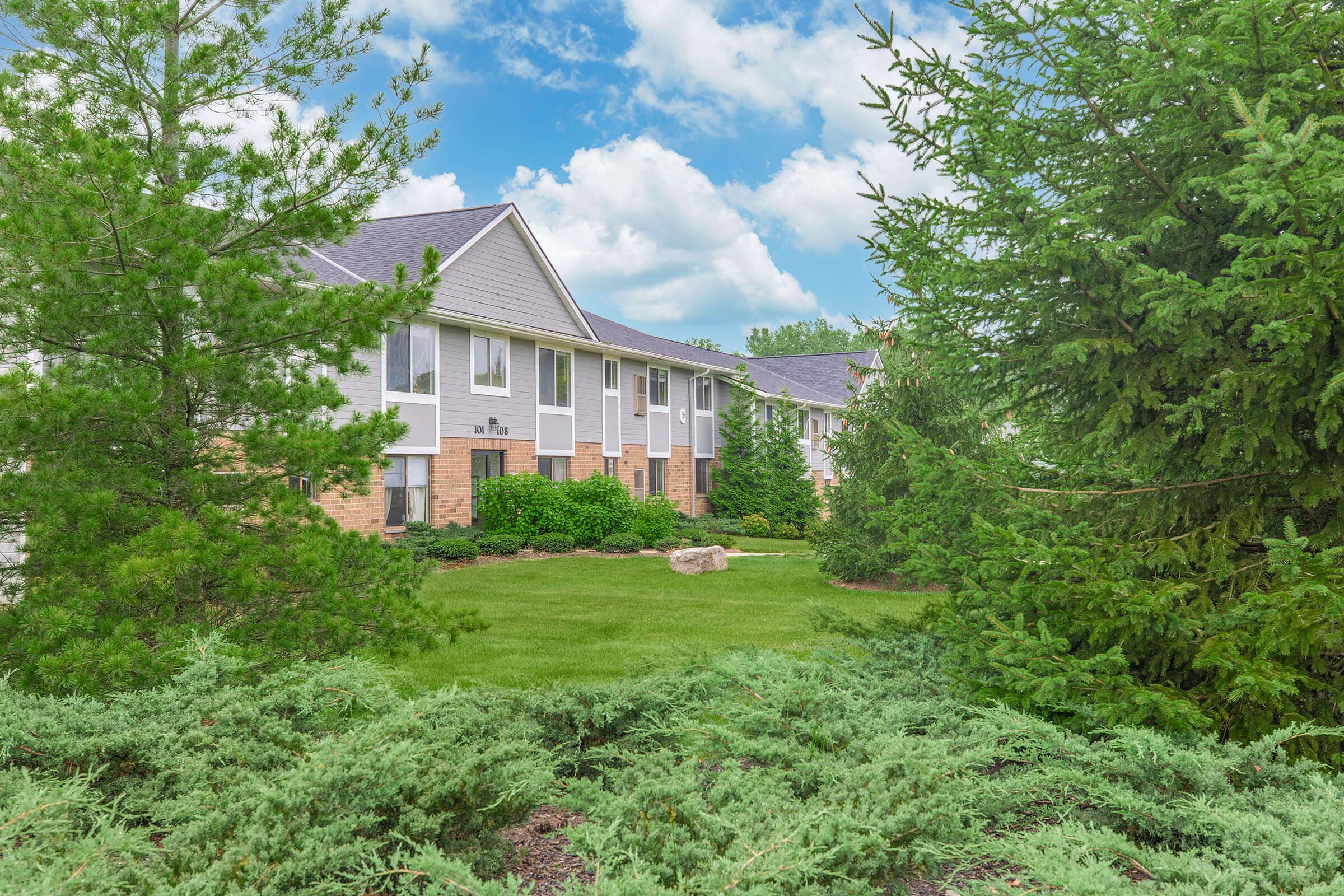 A well-maintained building surrounded by lush greenery, including tall evergreen trees and shrubs. The sky is partly cloudy, creating a bright atmosphere. The lawn is well-kept, featuring a rock, and the setting appears tranquil and inviting.