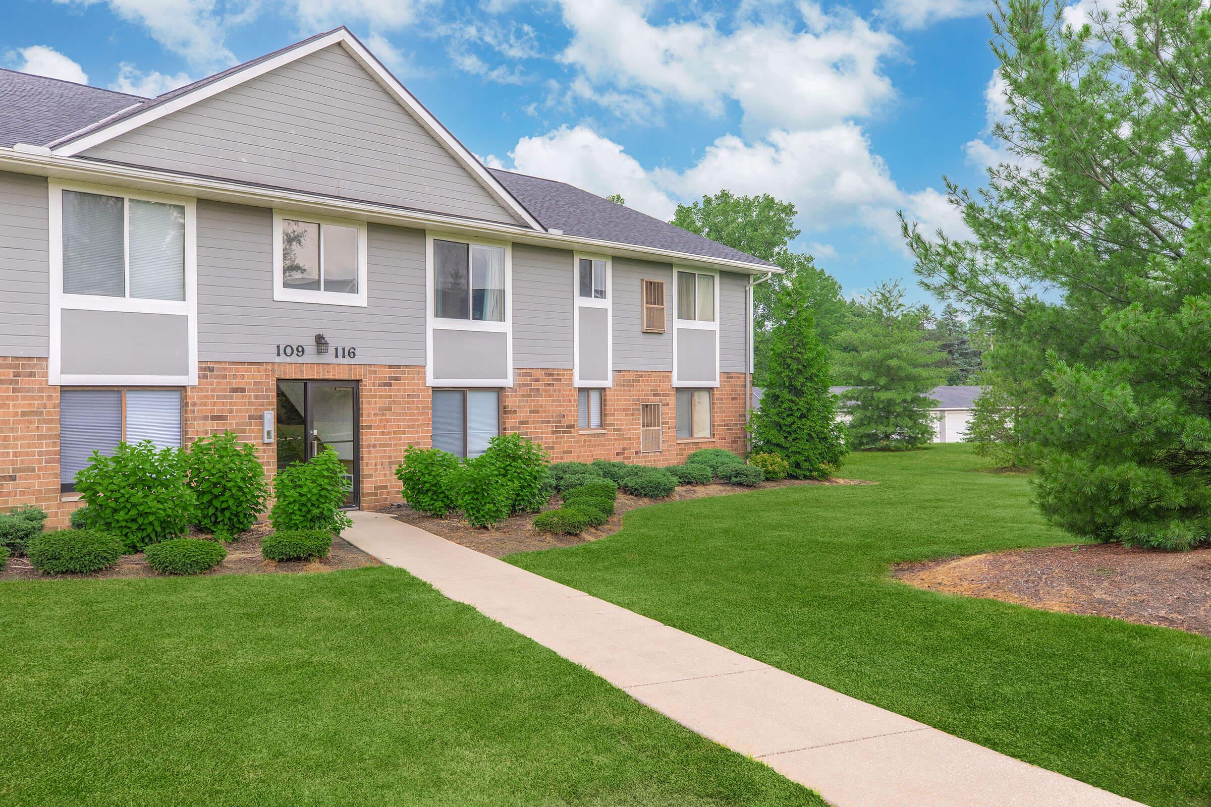 A well-maintained apartment building with a brick and siding exterior, surrounded by lush green lawns and ornamental shrubs. A paved walkway leads to the entrance, and trees are visible in the background under a blue sky with fluffy clouds.