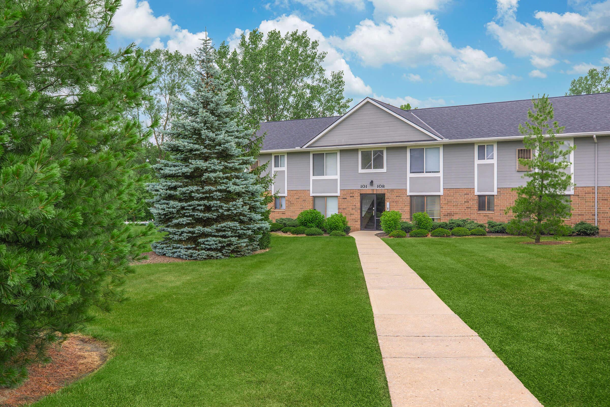 A well-maintained residential building with a landscaped front yard, featuring green lawns, shrubs, and trees. A paved walkway leads to the entrance, and the sky is partly cloudy, creating a pleasant atmosphere.