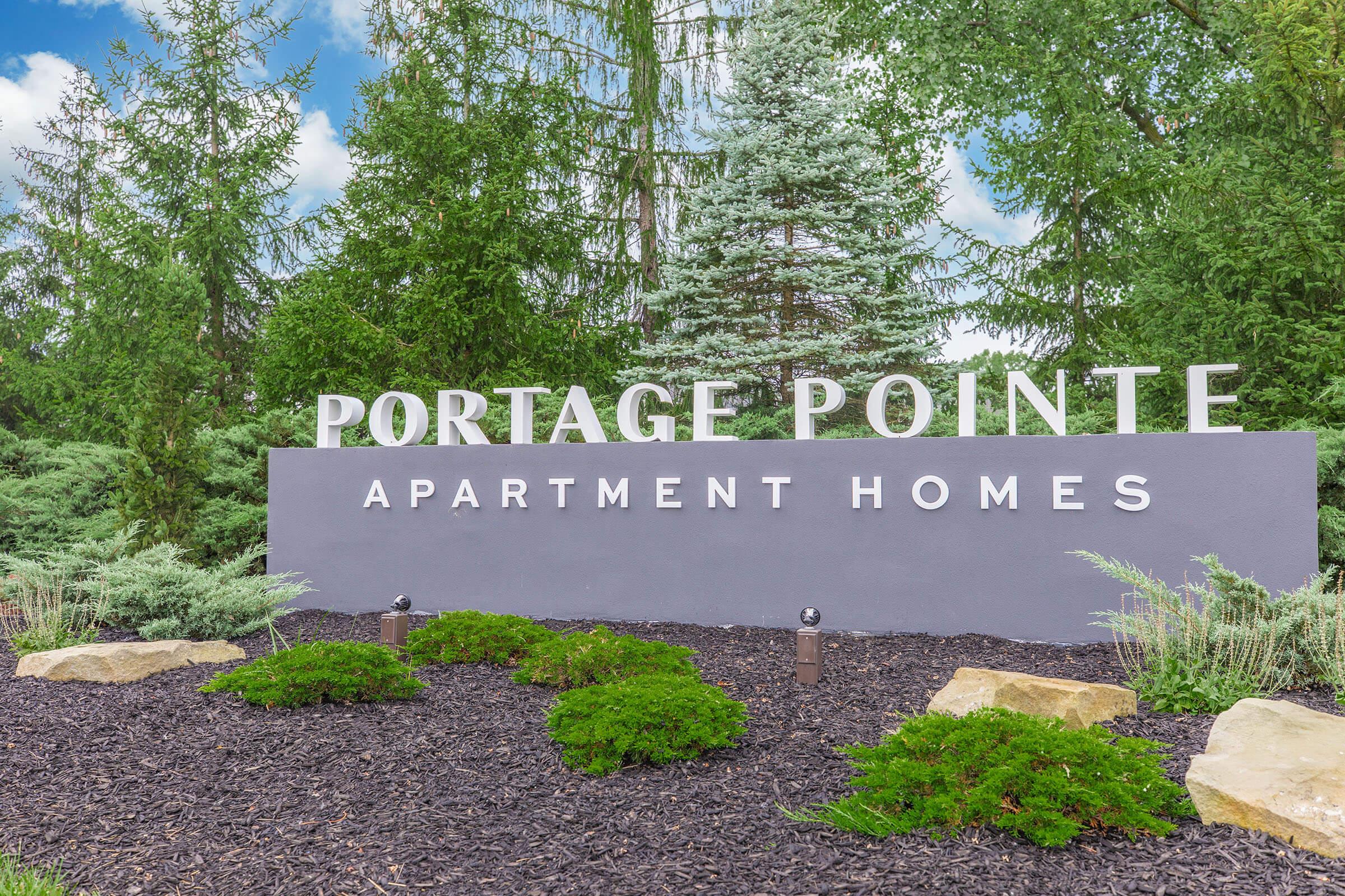 Sign for Portage Pointe Apartment Homes displayed prominently in front of a landscaped area featuring trees and shrubs. The sign has a modern design with the name "Portage Pointe" in large letters above "Apartment Homes" on a solid background, surrounded by decorative stones and greenery.