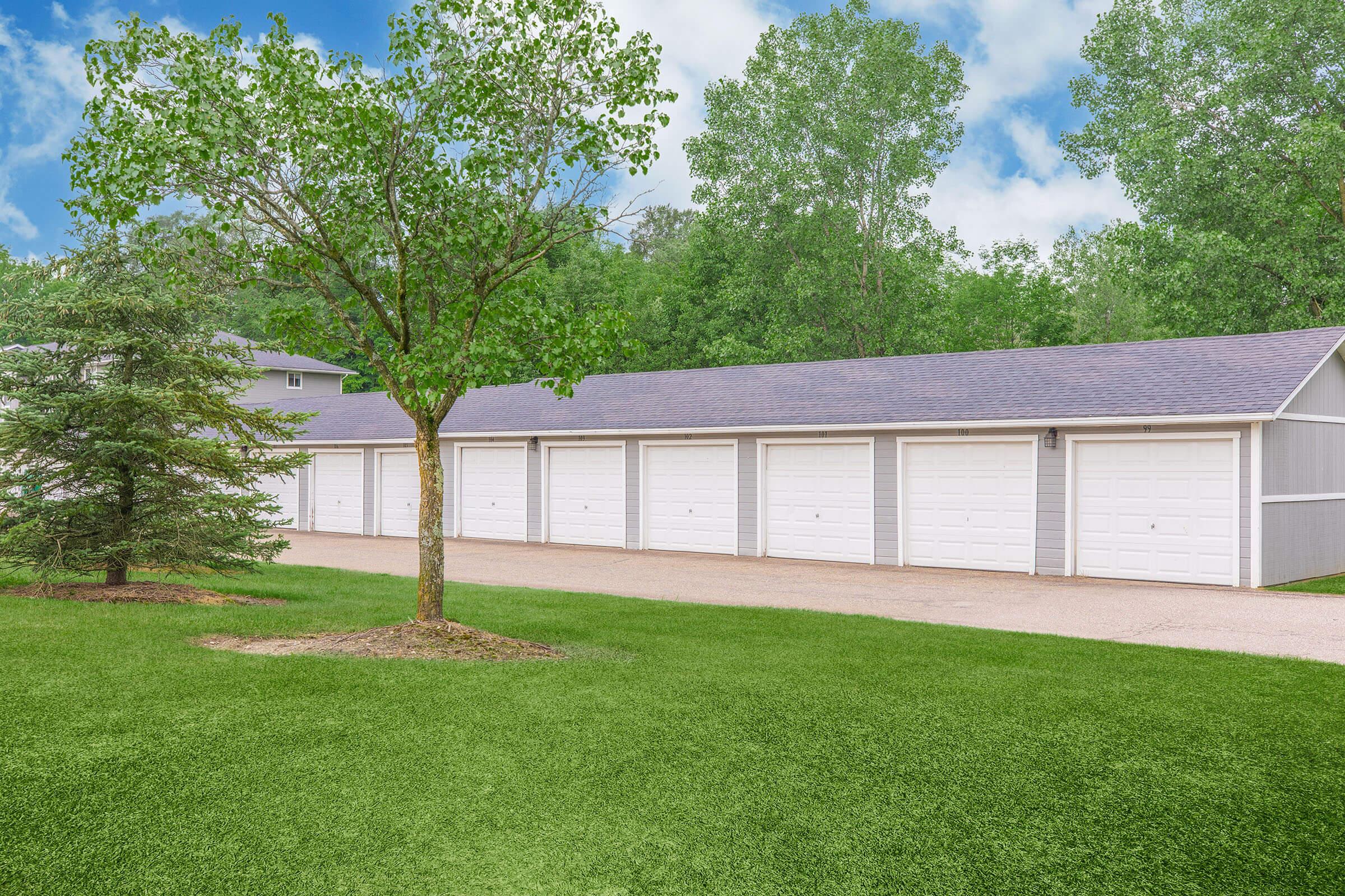 A row of metallic storage garages with white doors, set against a backdrop of green trees and a blue sky. A tree stands in the foreground, and a grassy area surrounds the garages, creating a serene outdoor environment.