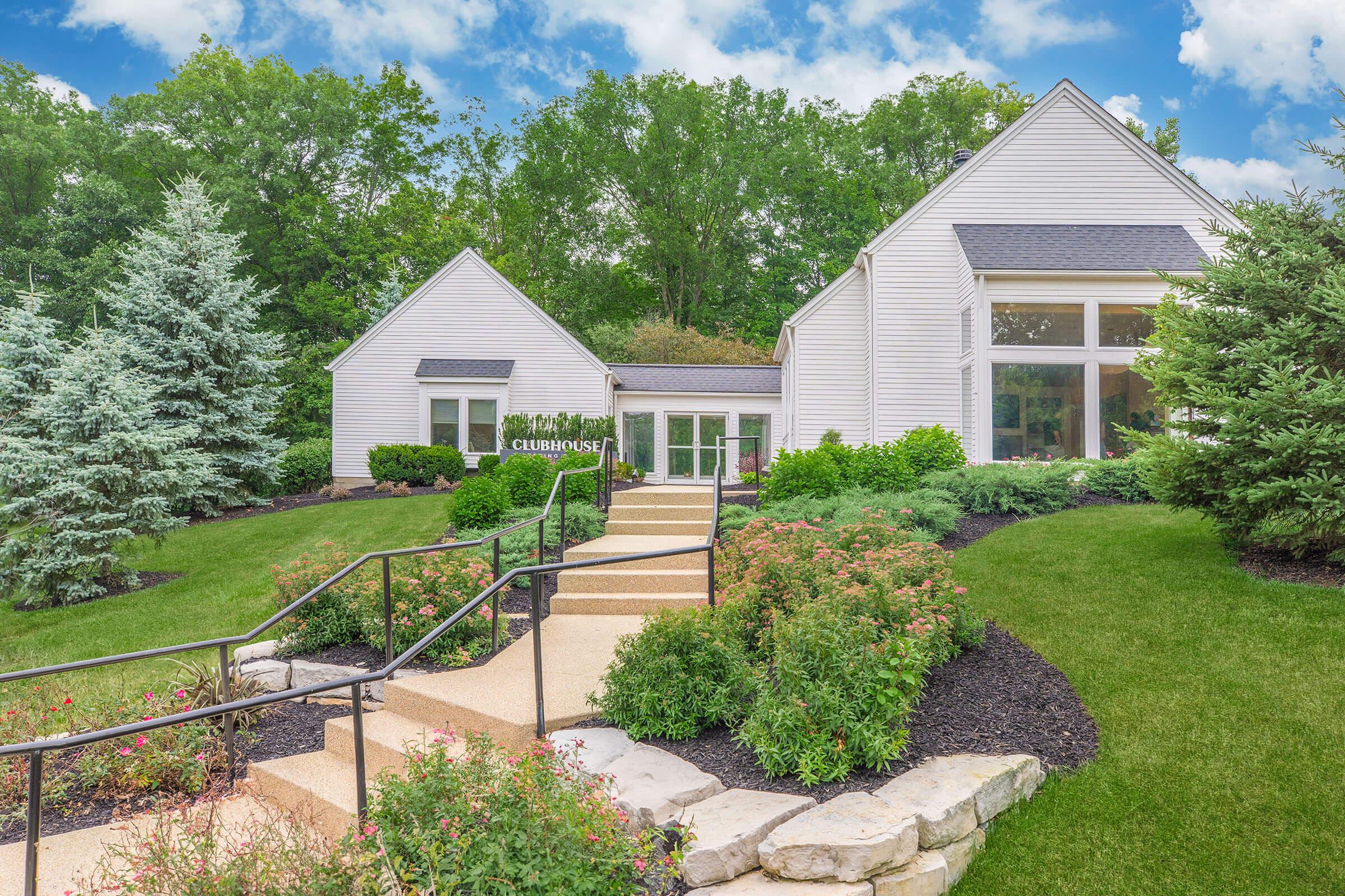 A well-maintained clubhouse surrounded by lush greenery, featuring a landscaped pathway with stone steps leading up to the entrance. Flanking the pathway are vibrant flower beds and tall trees, creating a serene and inviting atmosphere, under a bright blue sky with scattered clouds.
