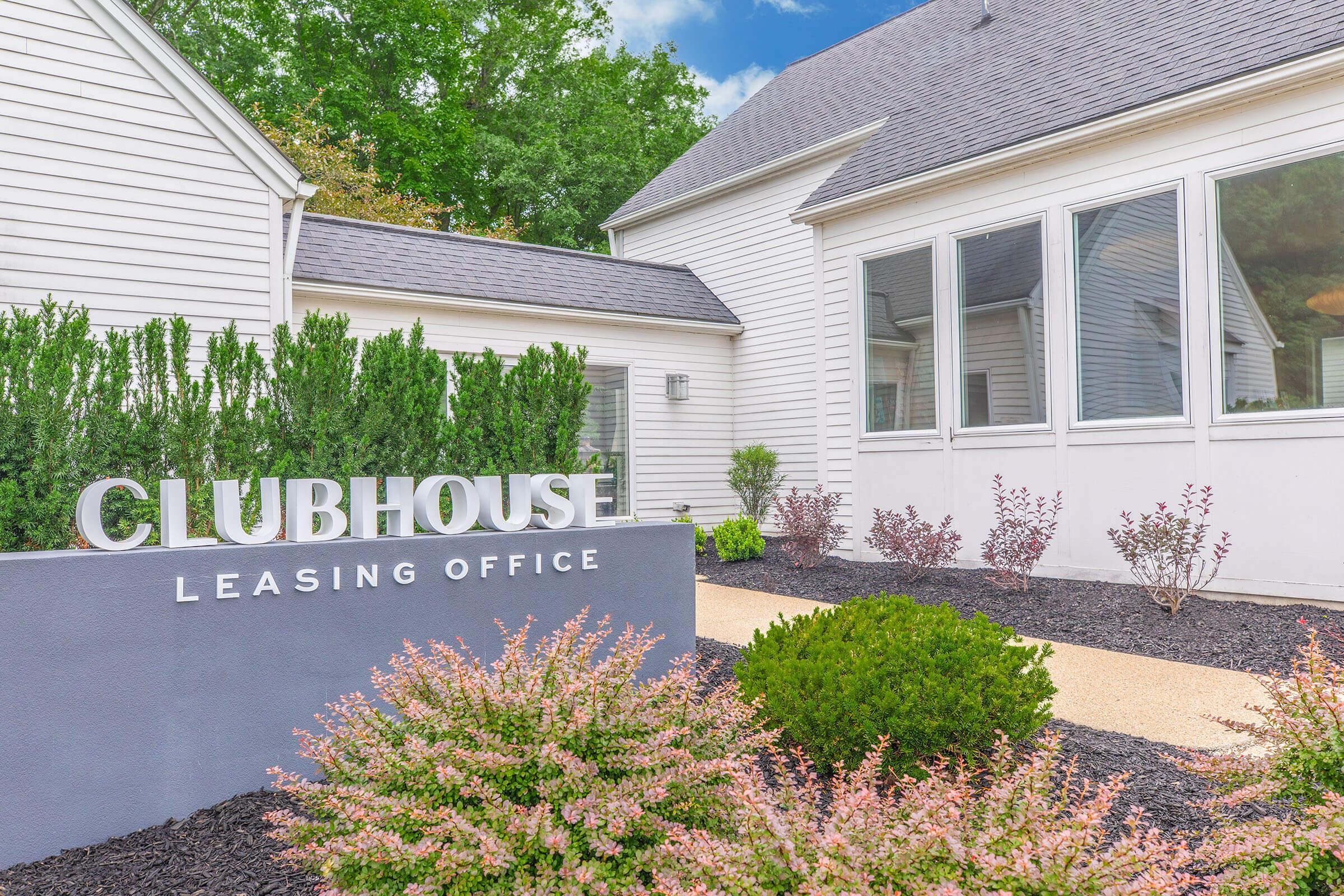 Sign for "Clubhouse Leasing Office" in a well-maintained outdoor area featuring decorative shrubs and flowers, a modern building with large windows, and a pathway leading to the entrance. Green trees and a blue sky create a welcoming atmosphere.