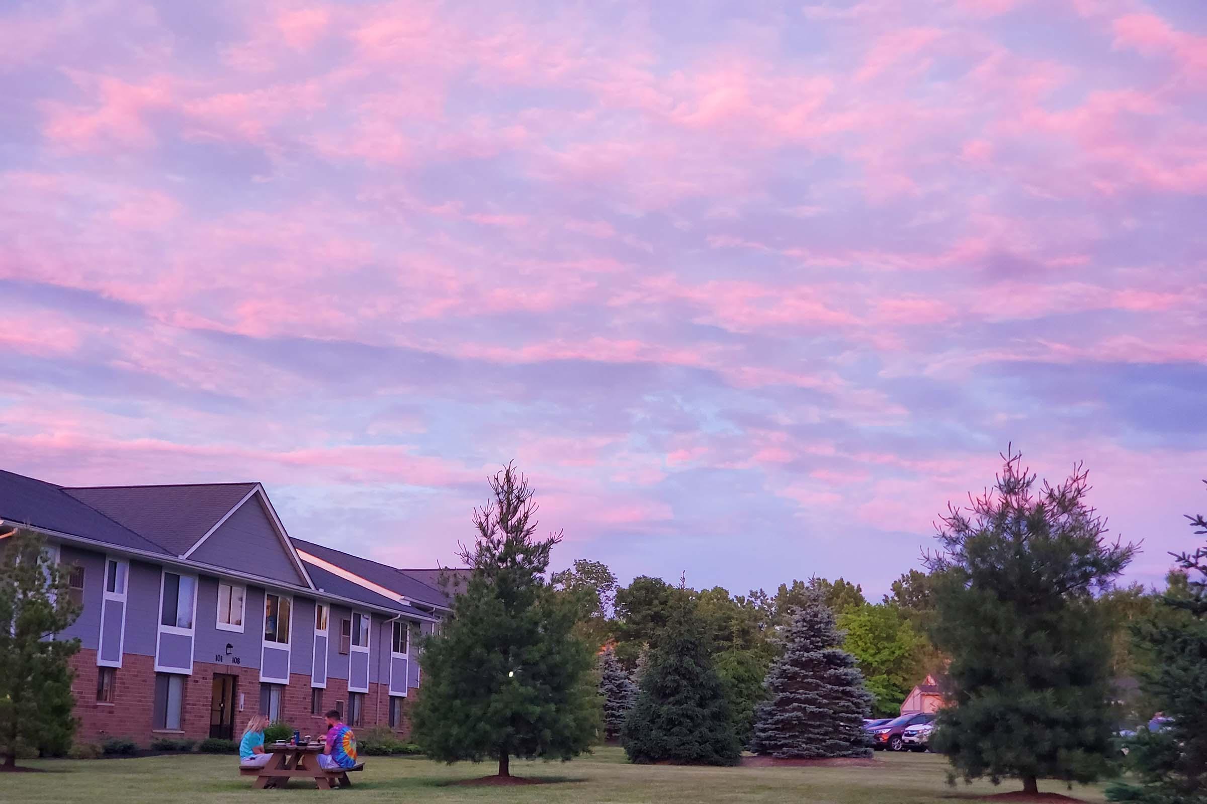 A serene outdoor scene featuring a colorful sunset sky with shades of pink and purple. In the foreground, two people are seated at a picnic table, while evergreen trees border the area. A building is visible on the left, along with a line of parked cars in the background.