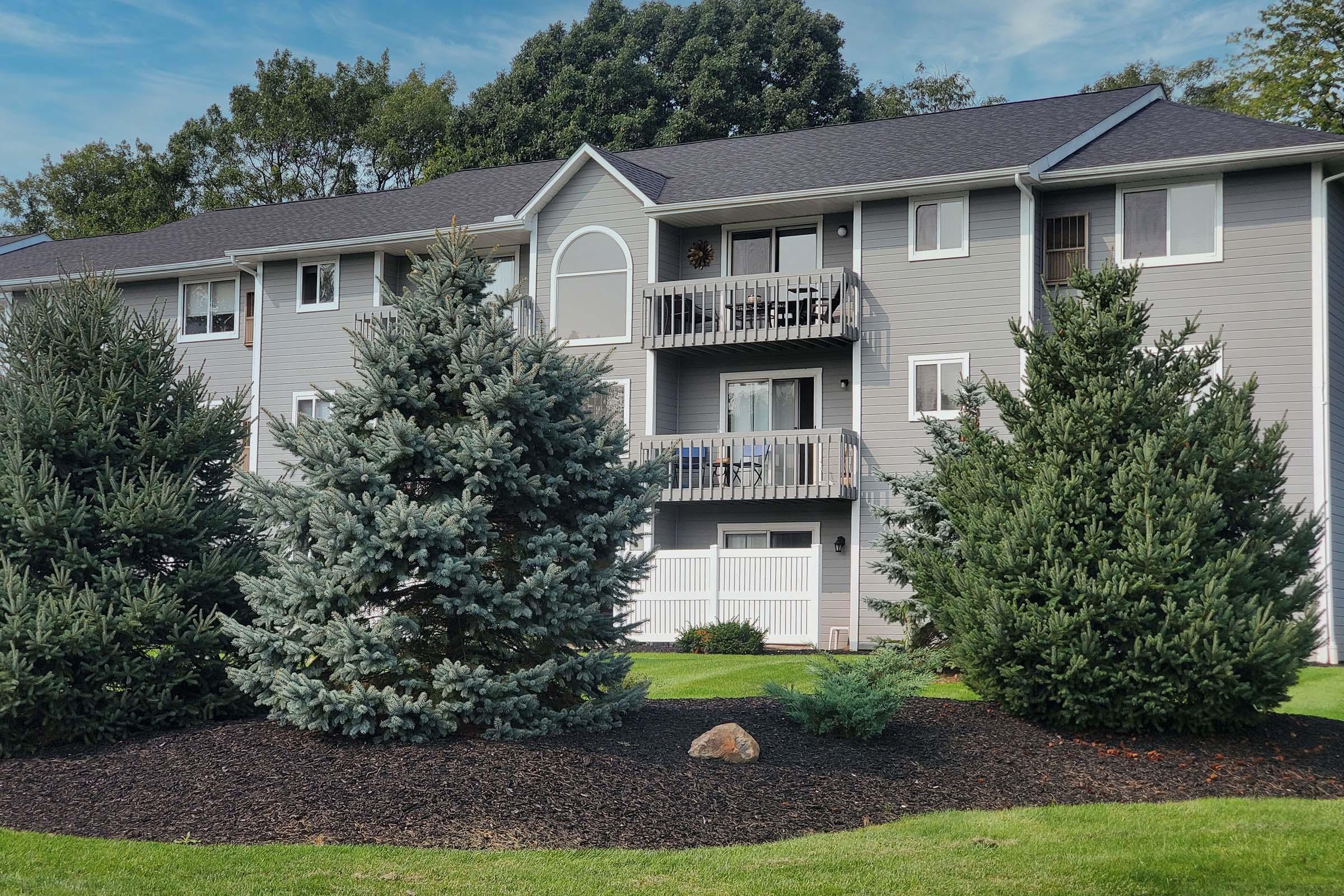 A modern multi-story apartment building with gray siding, featuring balconies and white railings. The foreground includes several evergreen trees and well-maintained landscaping with decorative rocks and mulch. The sky is partly cloudy, indicating a pleasant day.