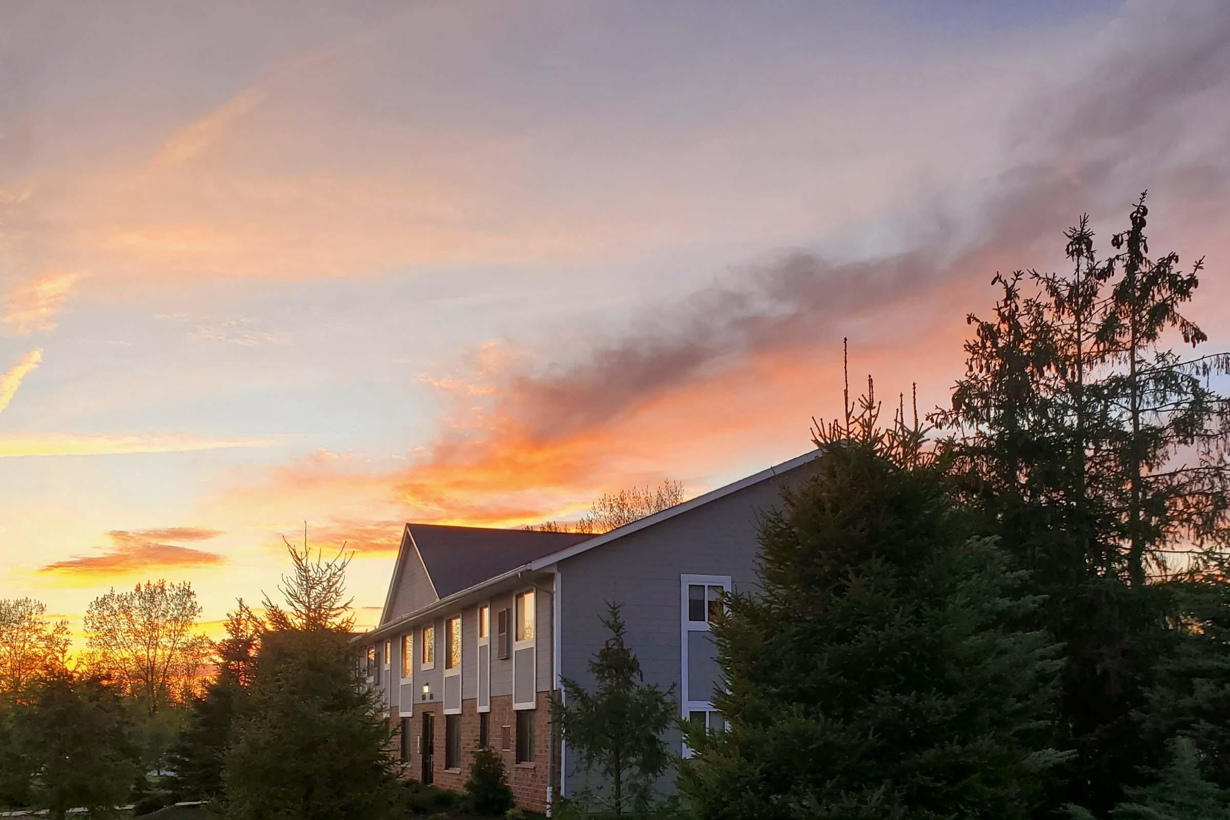 A scenic view of a building with a light-colored exterior and dark roof, surrounded by tall evergreen trees. The sky is painted with vibrant hues of orange, pink, and purple as the sun sets, creating a tranquil atmosphere. Wispy clouds are visible, enhancing the beauty of the sunset.
