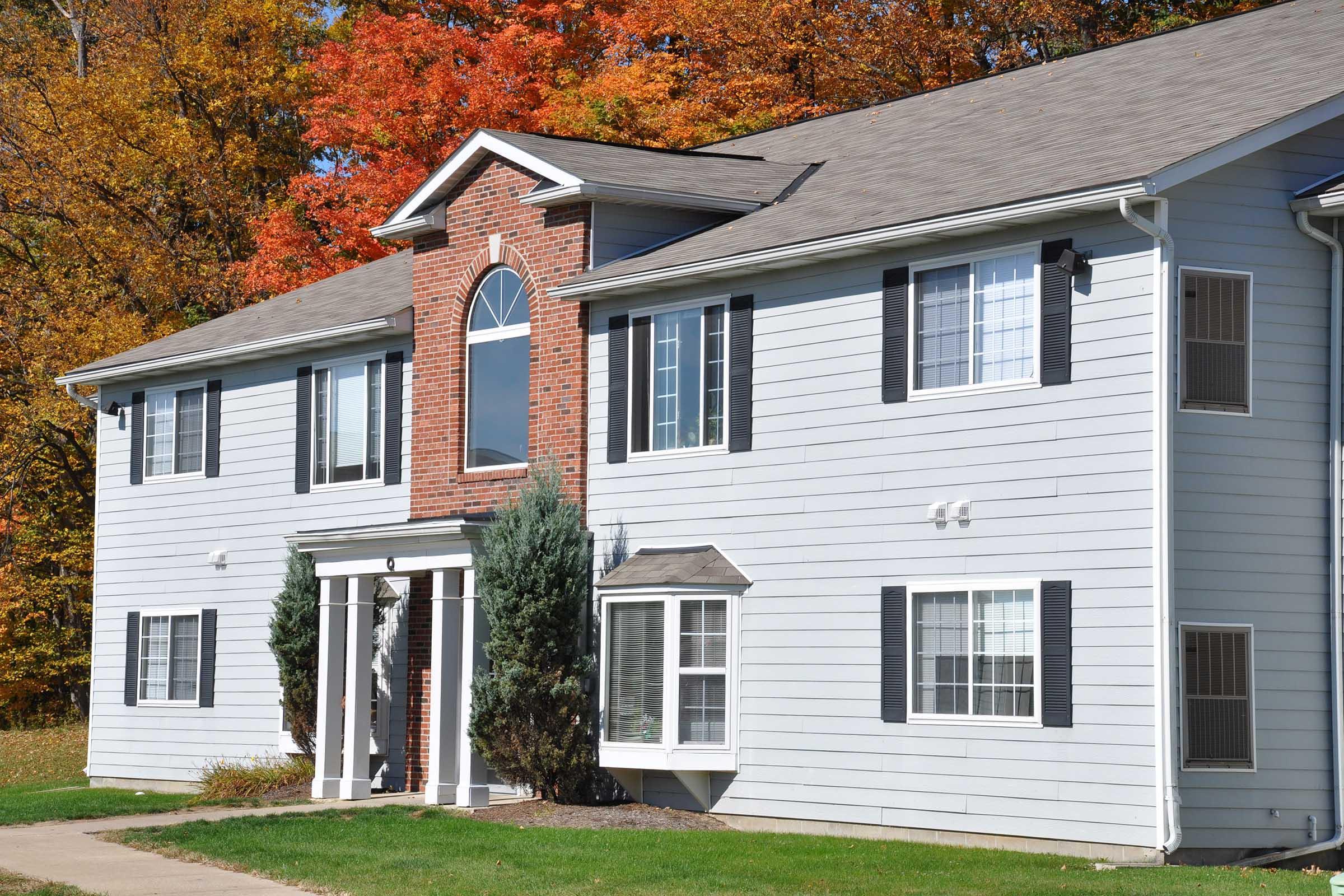 A two-story residential building with a mix of light gray and brick exterior. It features multiple windows with shutters, a prominent arched window at the top center, and well-maintained landscaping. In the background, vibrant autumn trees display a variety of orange and yellow foliage.