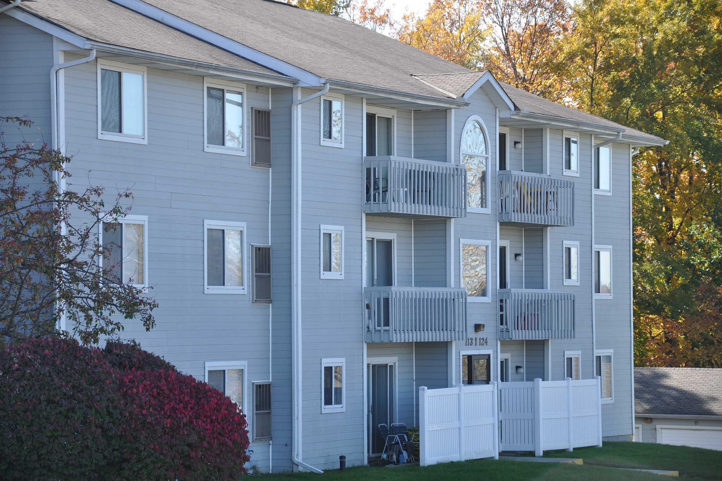 Gray multi-unit residential building with balconies, surrounded by trees showing autumn foliage. A white picket fence is in front, and a small lawn is visible.