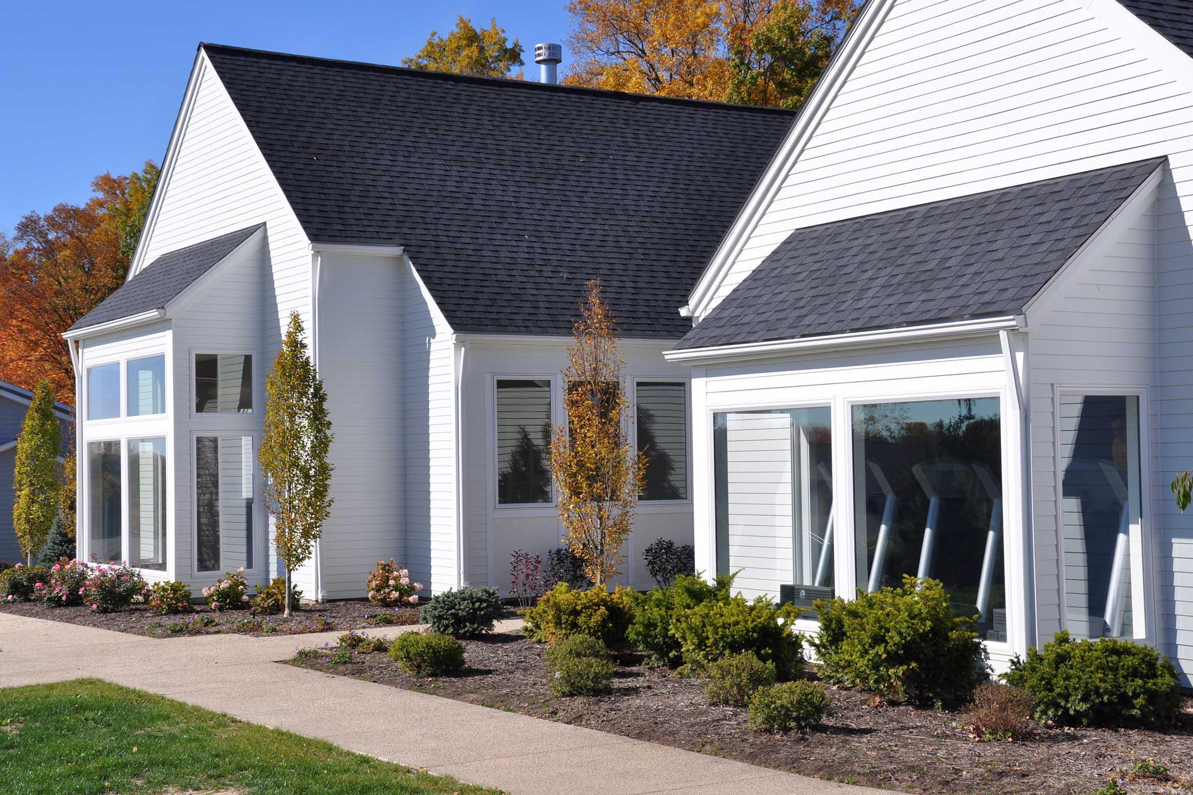 A modern white house with a peaked roof, featuring large windows and a landscaped front yard. The yard has neatly trimmed shrubs and flower beds, with vibrant autumn foliage in the background. The scene is bright and sunny, highlighting the home's architecture and surrounding greenery.