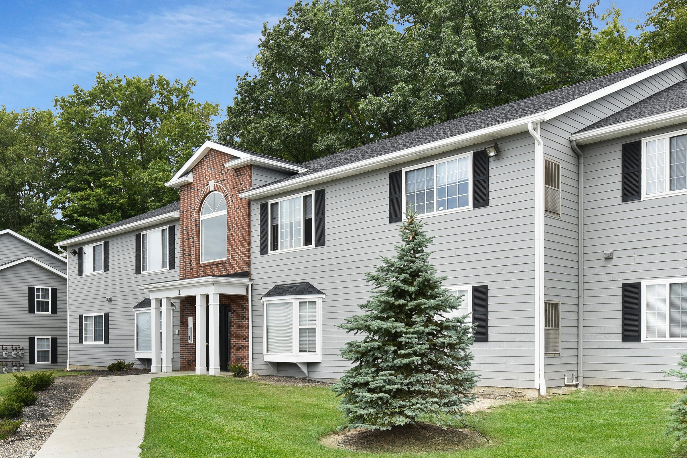 Two-story residential building with gray siding and brick accents, featuring white windows and black shutters. A neatly maintained lawn with a small evergreen tree is in the foreground, and a walkway leads to the entrance. Trees line the background, suggesting a suburban setting.