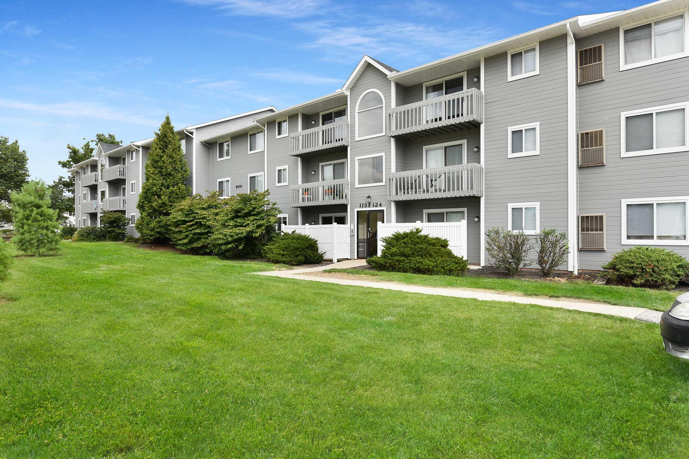A multi-story apartment building with gray siding, multiple balconies, and a well-maintained lawn. The scene features a clear blue sky and manicured shrubs, providing a welcoming residential environment. A pathway leads to the entrance of the building.