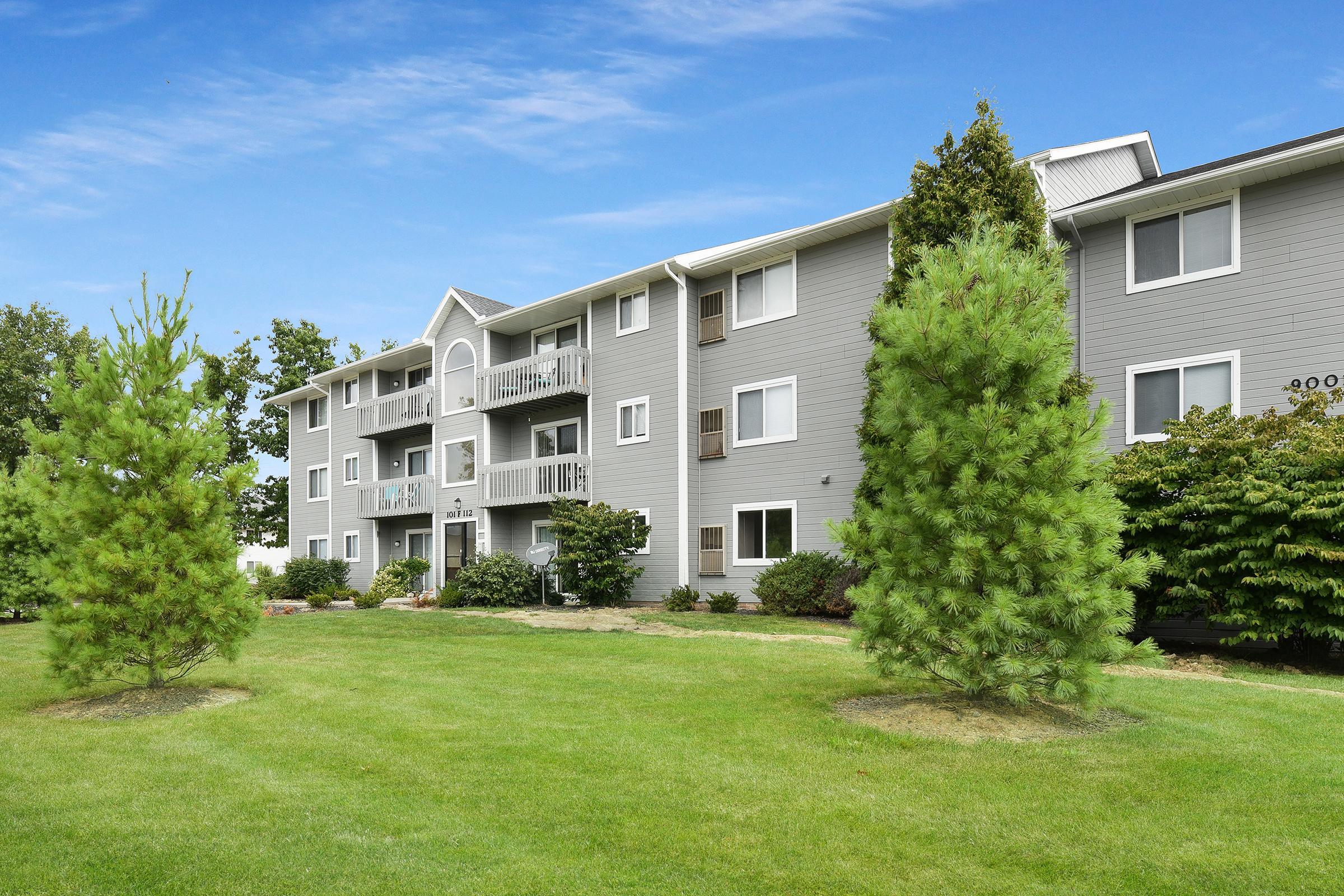 A multi-story gray apartment building surrounded by green lawns and trees. The building features balconies on the upper floors, with neatly manicured landscaping in front. The sky is clear and blue, adding to the inviting atmosphere of the residential area.