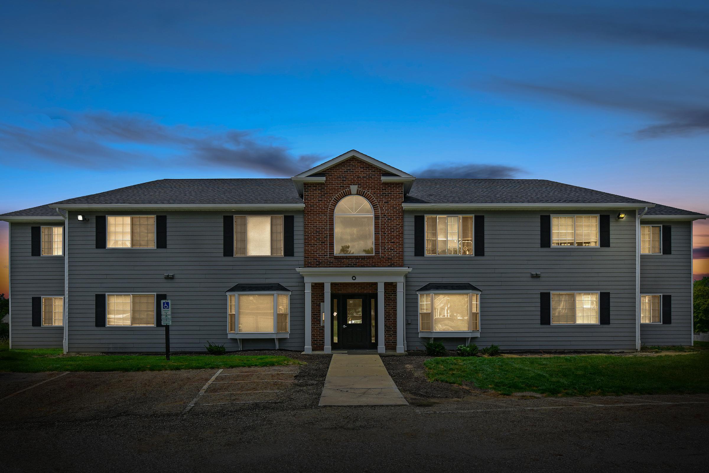 A two-story residential building with a grey exterior and brick accents. The front features large windows, some illuminated, and a central arched window above the entrance. The setting is at dusk with a blue sky and soft clouds. A small parking lot is visible in front of the building.