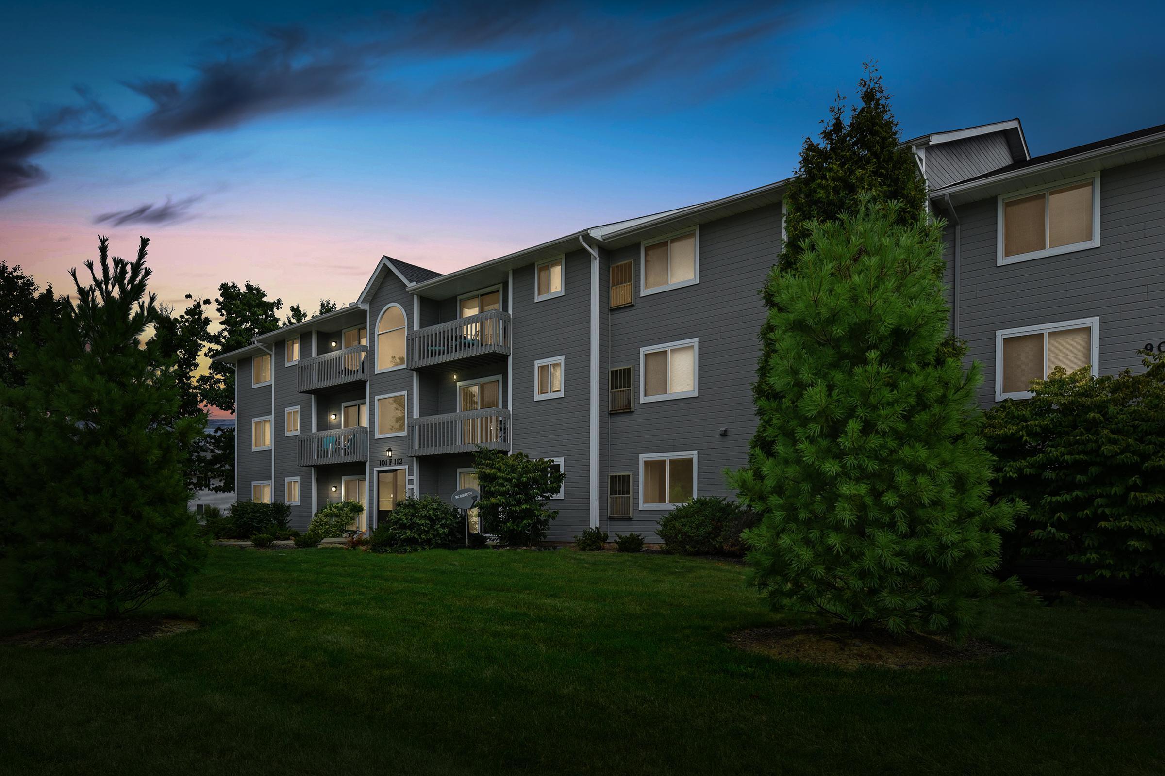A cozy apartment building with multiple balconies, surrounded by green trees and well-maintained landscaping. The sky displays a gradient of sunset colors, creating a warm and inviting atmosphere. The scene captures the essence of suburban living in a peaceful setting.