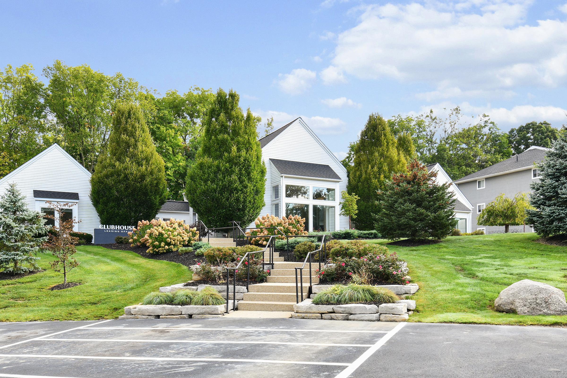 A landscaped entrance to a community with well-maintained greenery, flowering bushes, and trees. There are stone steps leading to a modern building, with a parking area in the foreground. The sky is blue with scattered clouds, creating a welcoming atmosphere.