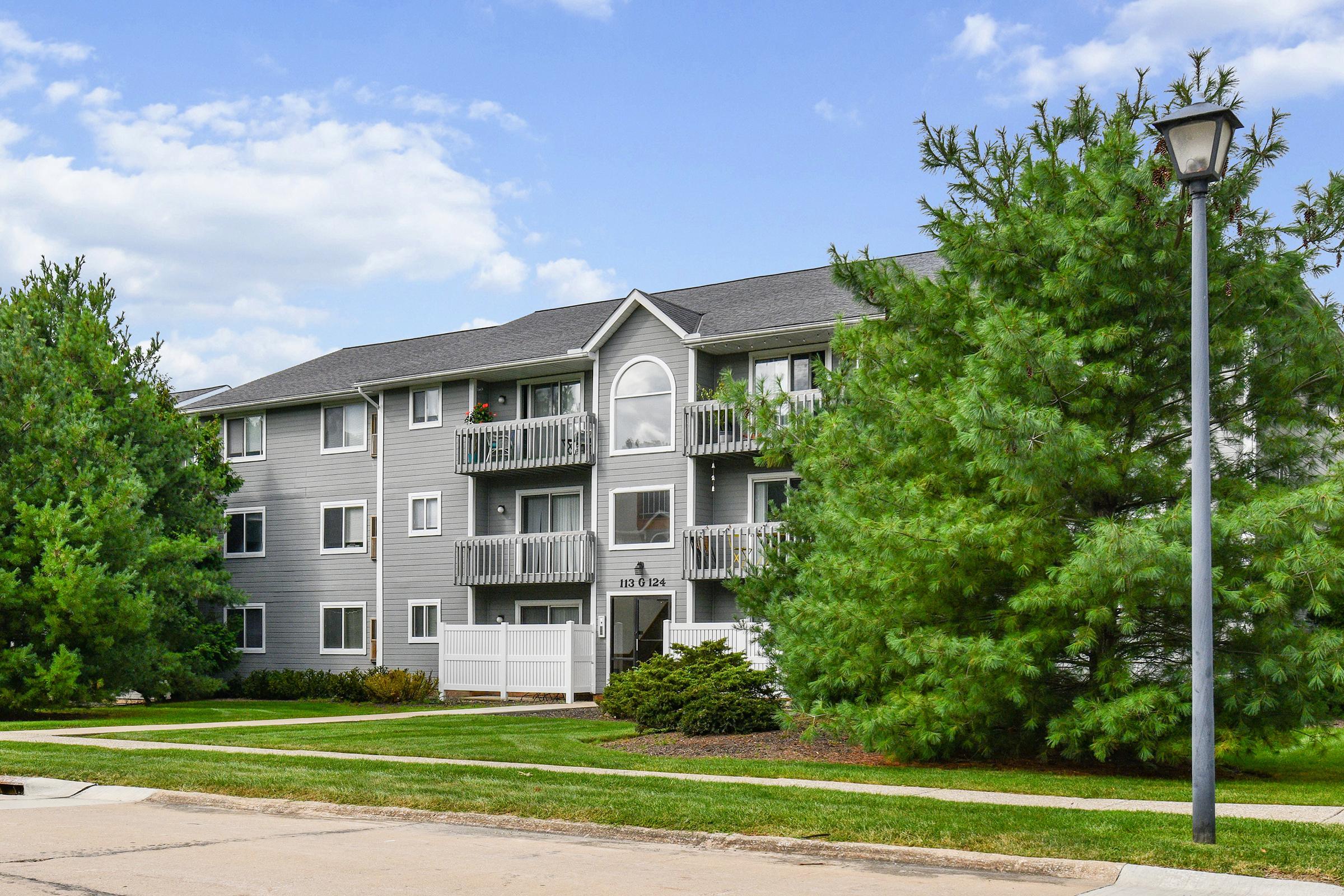 A multi-story apartment building with a gray exterior, featuring balconies and decorative arches. Surrounding the building are lush green trees and well-maintained landscaping. A concrete path leads to the entrance, and a street lamp stands nearby under a partly cloudy sky.