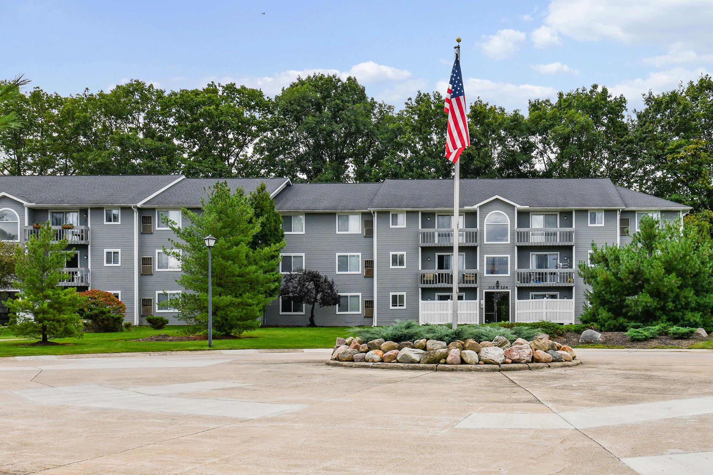 A multi-story residential apartment building with gray siding, surrounded by green trees. In the foreground, a circular stone arrangement and an American flag on a pole are prominently displayed. The sky is partly cloudy, creating a pleasant atmosphere.