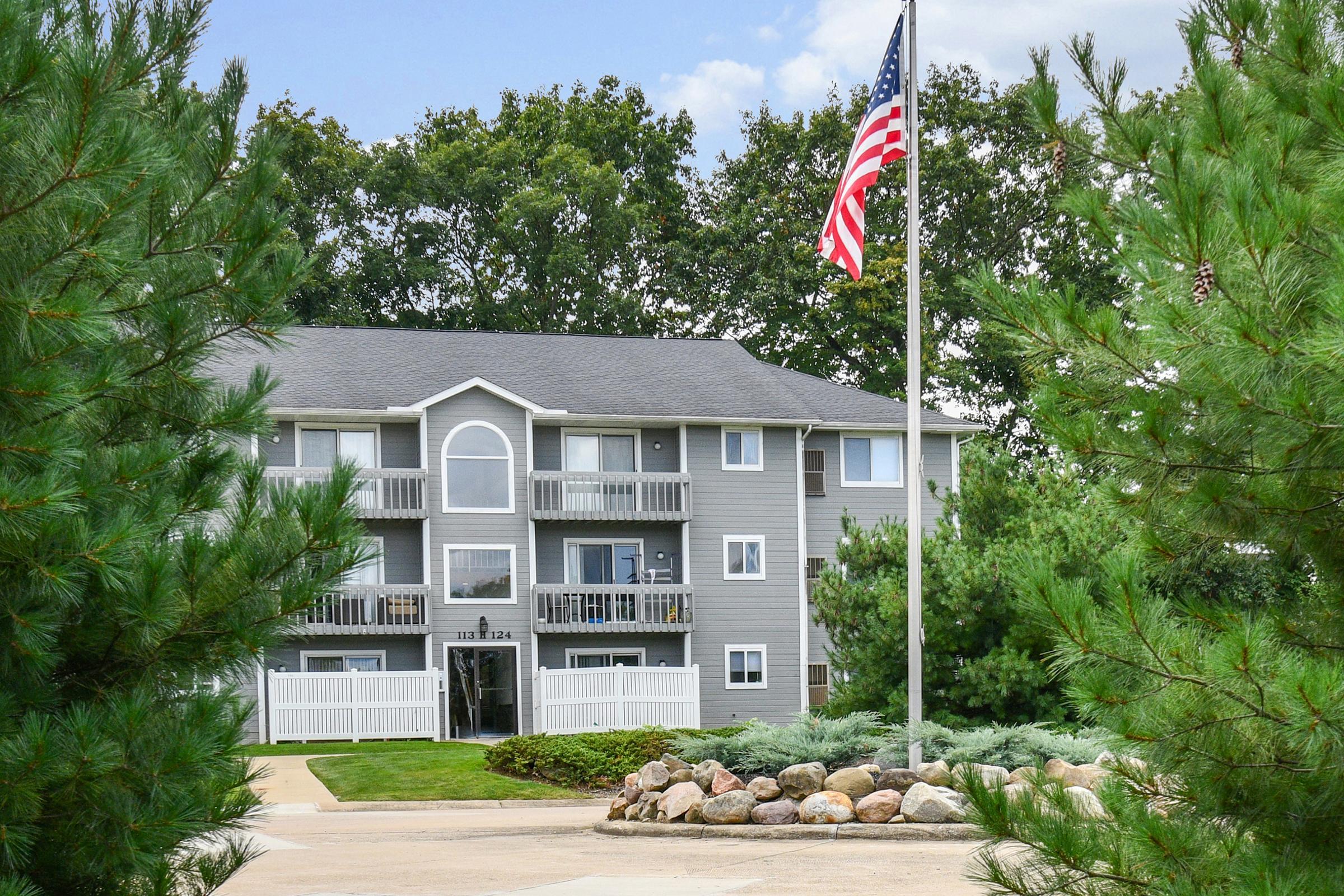 A three-story gray apartment building with white balconies, surrounded by greenery and tall pine trees. An American flag stands on a pole in front of the building, and a landscaped area with rocks and shrubs is visible along the driveway. The sky is partly cloudy.