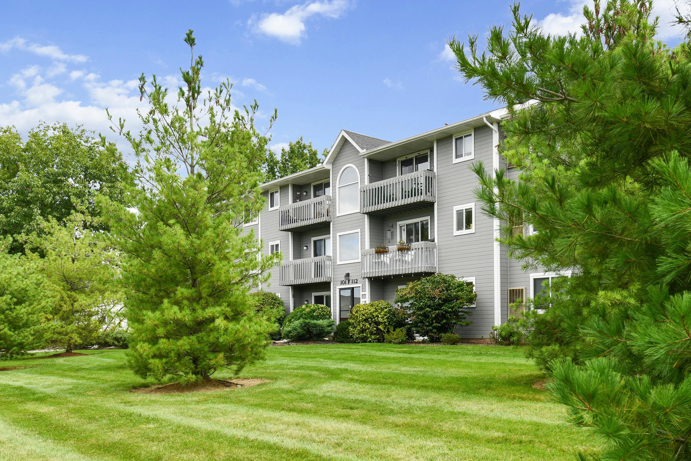 A three-story residential building with balconies set among lush greenery and well-maintained lawns. The scene features several tall trees and a clear blue sky, creating a peaceful suburban atmosphere.