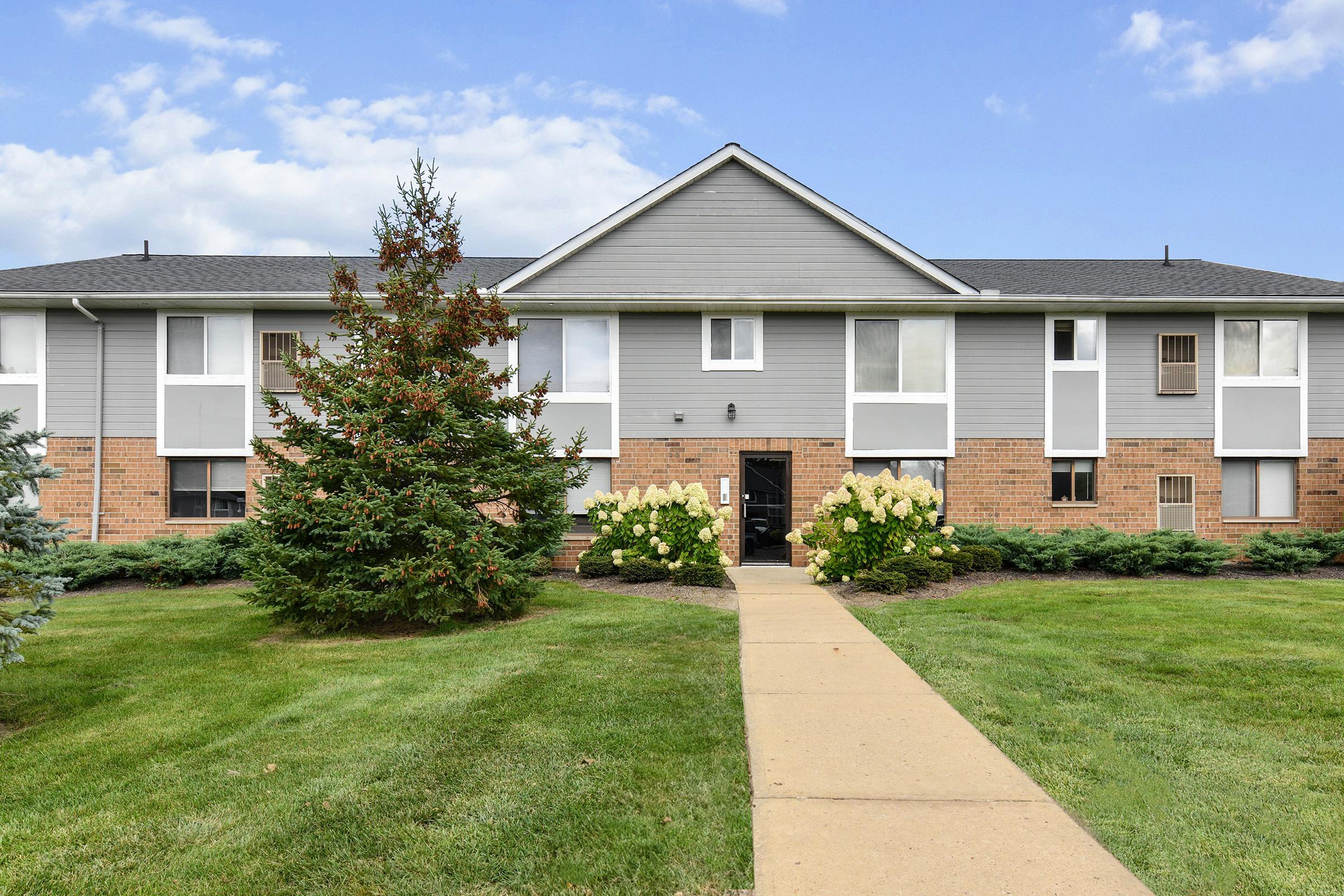 Exterior view of a two-story apartment building with gray siding and brick accents. Lush green lawns surround the building, featuring decorative landscaping with flowers and shrubs. A concrete walkway leads to the entrance, which has a clear view of the blue sky above.