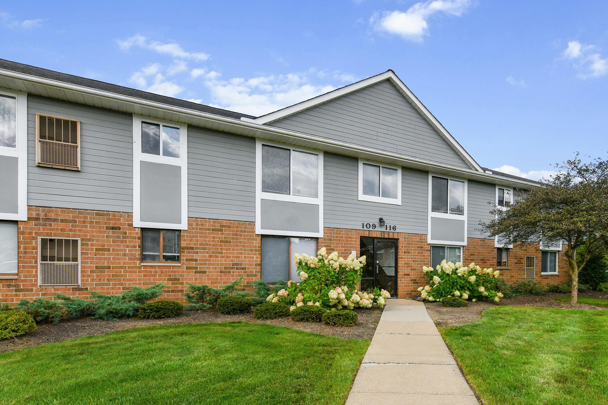 Low-rise apartment building with a mix of gray siding and brick exterior. The entrance features large windows and is surrounded by well-maintained landscaping, including green grass, shrubs, and hydrangea plants. The sky is partly cloudy, highlighting a pleasant day.