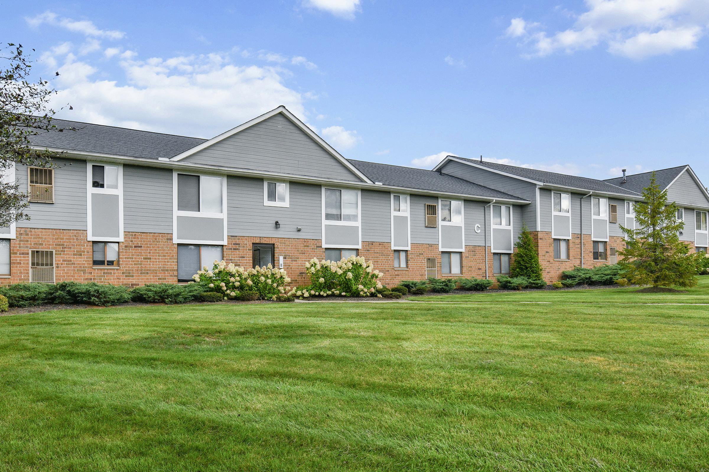 A low-rise apartment building with gray siding and brick accents, surrounded by lush green grass and landscaped areas. The building features multiple entrances and large windows under a clear blue sky with some clouds.