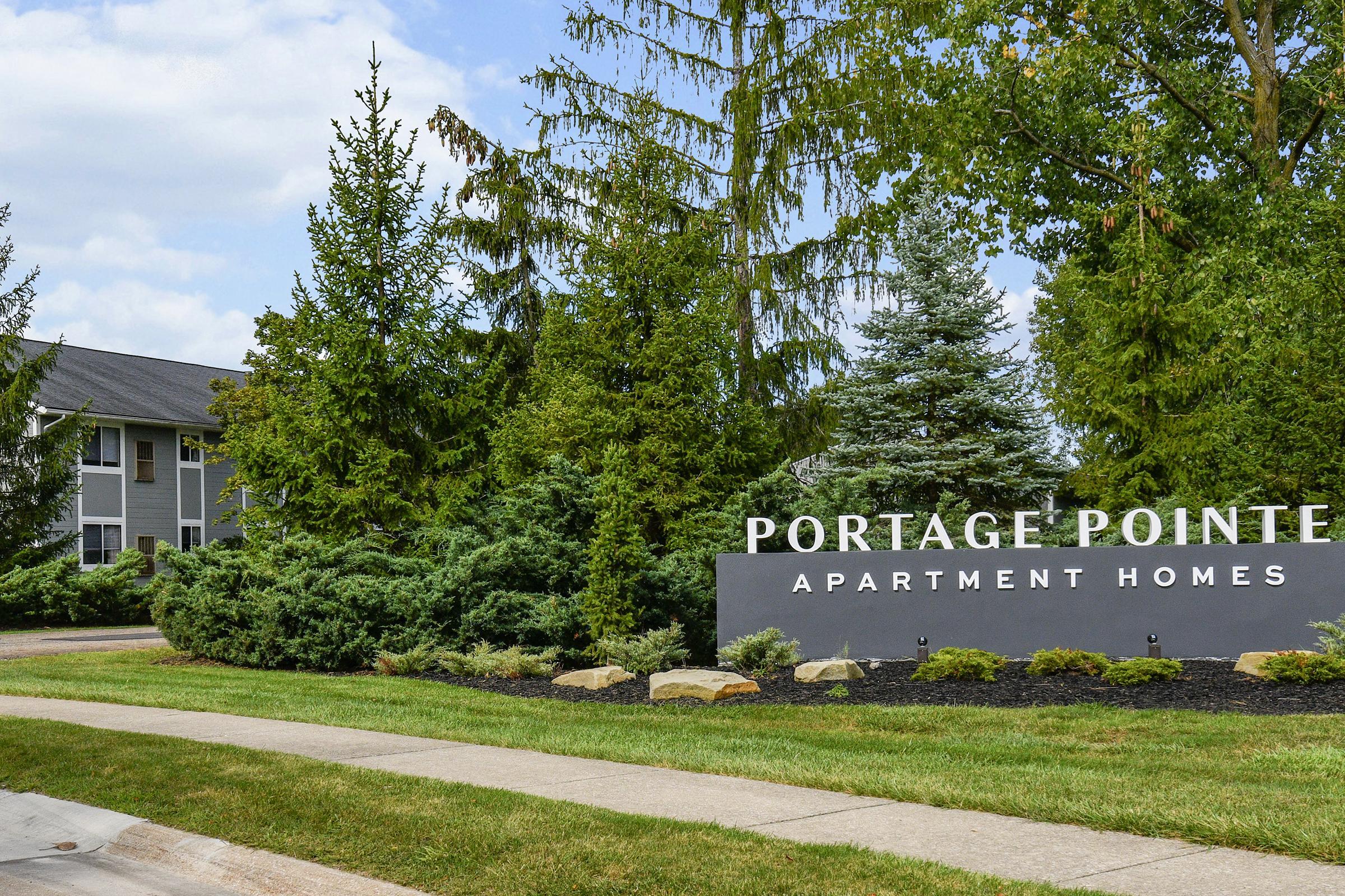 Sign for Portage Pointe Apartment Homes with well-maintained landscaping, including various trees and shrubs. A pathway leads up to the entrance, and a multi-story building is visible in the background under a partly cloudy sky.