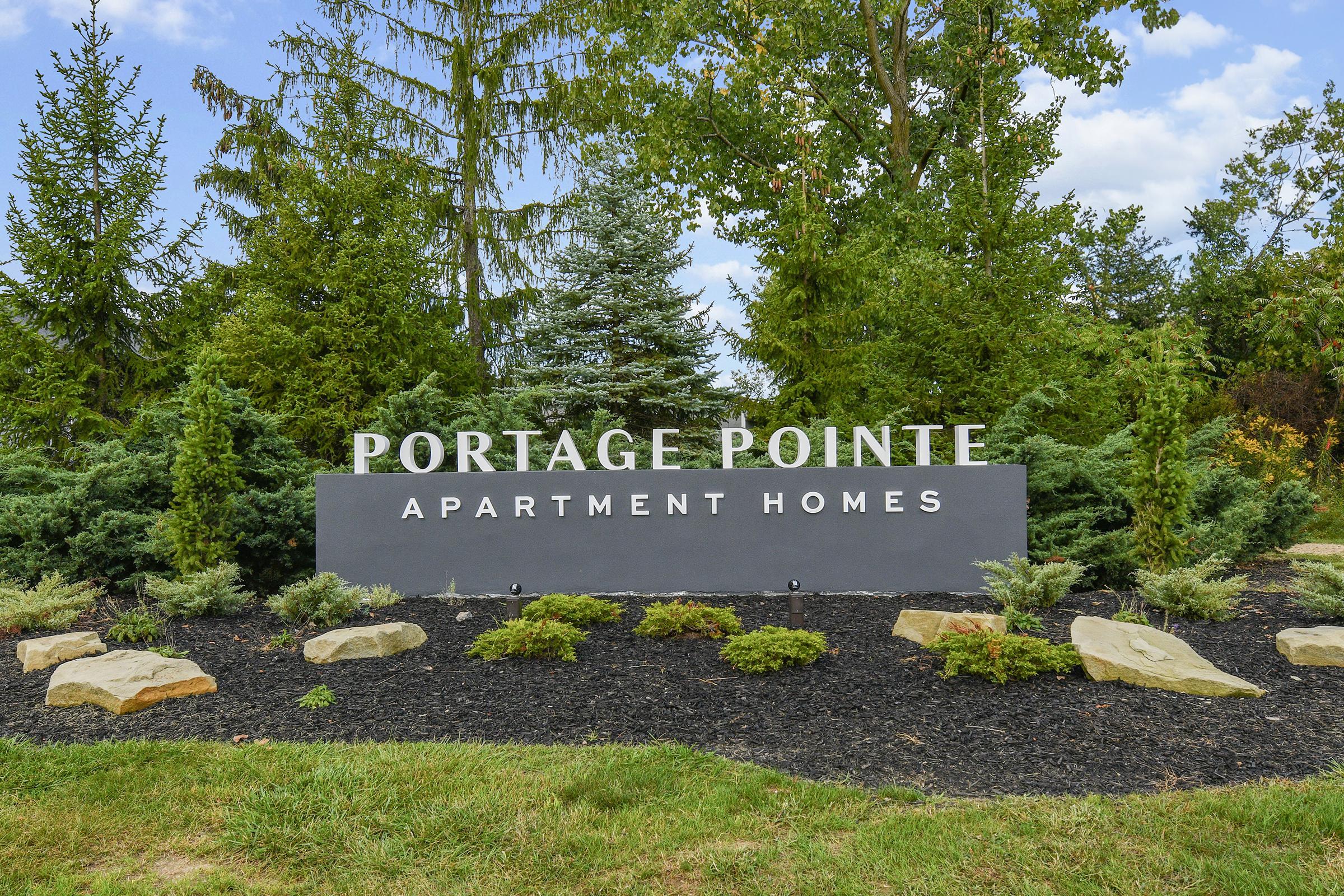 Sign for Portage Pointe Apartment Homes, surrounded by lush green trees and well-maintained landscaping featuring shrubs and rocks. The sign is prominently displayed in front of the apartment complex, highlighting the name of the residences.