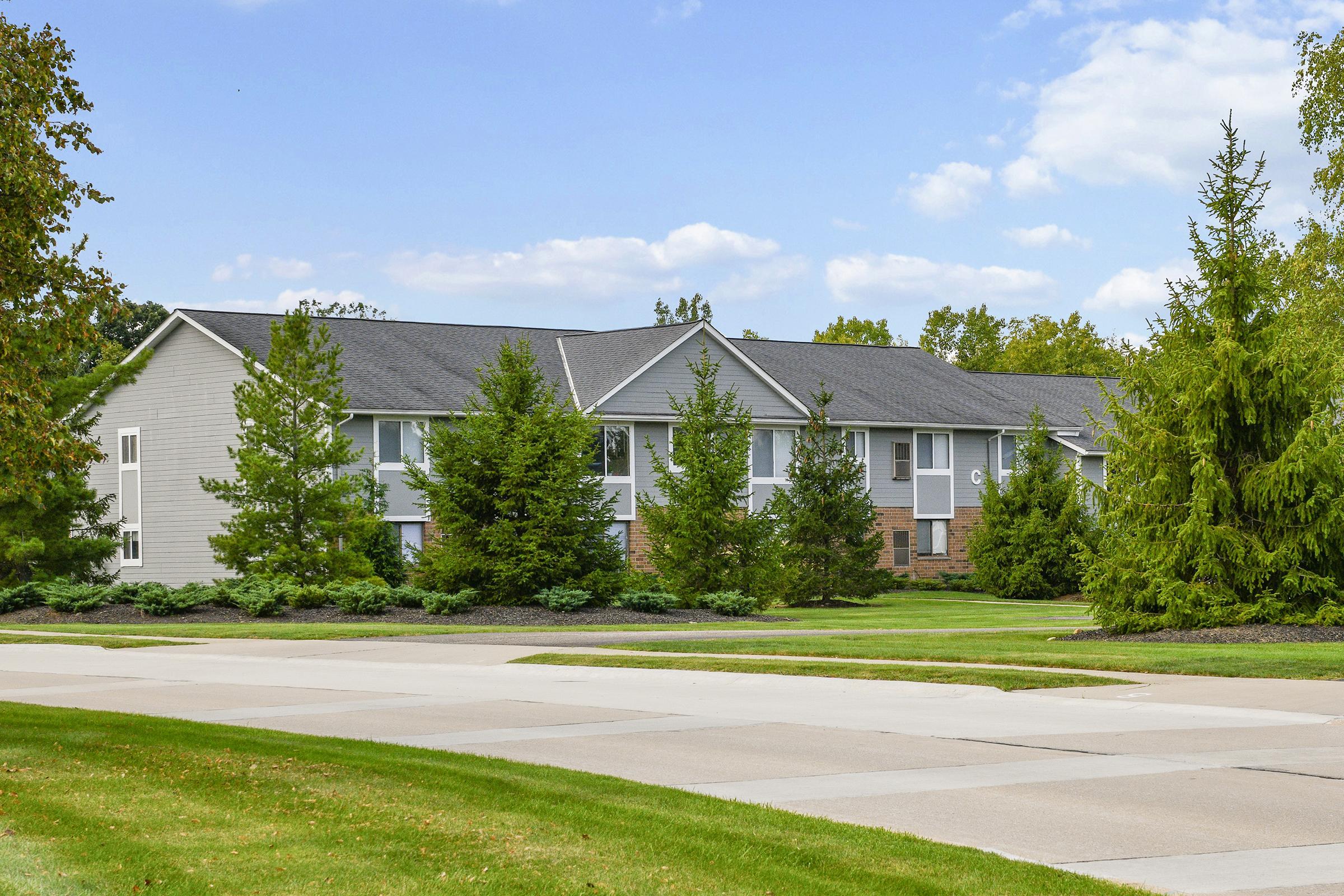 A modern multi-unit residential building with a gray exterior, surrounded by lush green trees and shrubs. A wide paved pathway leads to the building, and the sky is partly cloudy, adding to the inviting atmosphere of the neighborhood.