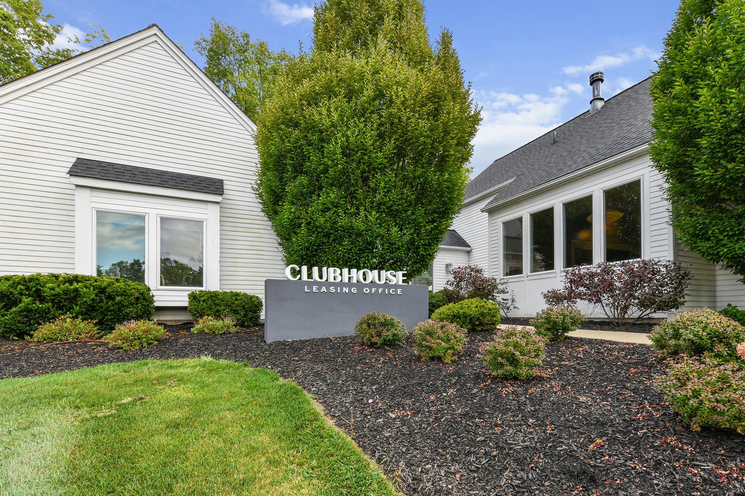 Sign for "Clubhouse Leasing Office" in front of a well-maintained building, surrounded by neatly trimmed bushes and landscaping, with a sunny sky in the background. The office features large windows, and the pathway is lined with dark mulch and small shrubs.
