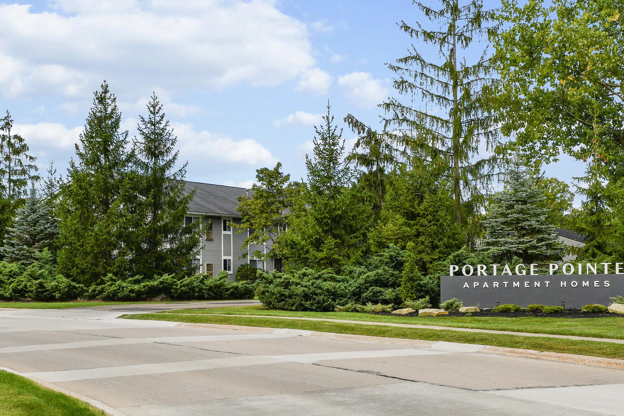 Sign for Portage Pointe Apartment Homes at the entrance, surrounded by well-maintained greenery and trees. A paved road leads into the apartment complex, with a clear blue sky in the background.