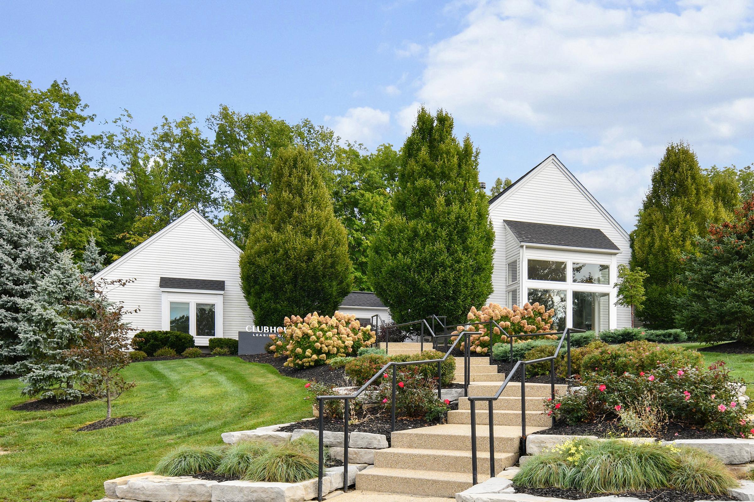 Two modern white buildings with large windows are set amidst lush greenery and neatly landscaped gardens. Stone steps lead up to the entrance, flanked by flowering bushes and trees. The sky is partly cloudy, adding a serene atmosphere to the scene.