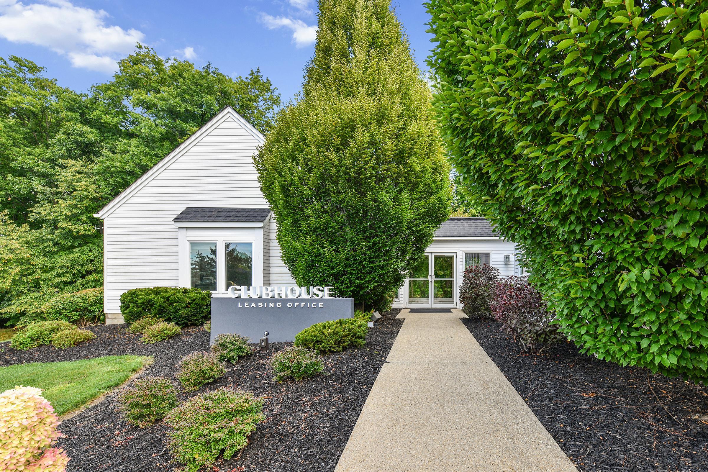 Exterior view of a leasing office with a modern sign displaying "CLUBHOUSE" and "LEASING OFFICE." The building features a white façade, surrounded by lush green landscaping, including tall trees and colorful shrubs. A paved pathway leads to the entrance, set against a clear blue sky.