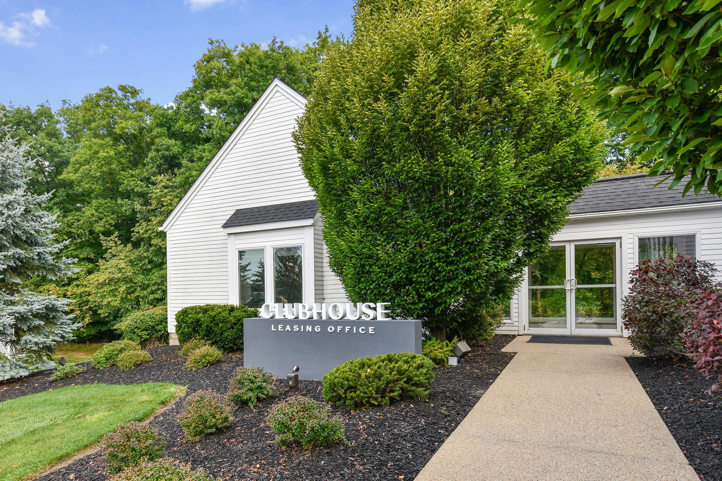 A white, multi-story building with a sloped roof and large front windows surrounded by greenery. A well-maintained pathway leads to the entrance, which features a sign that reads "Clubhouse Leasing Office," along with neatly trimmed shrubs and landscaping.