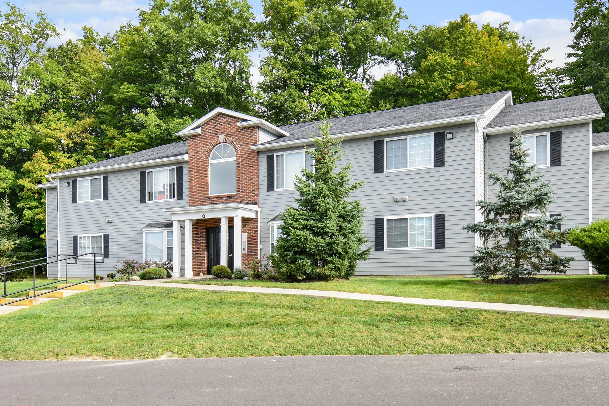A two-story apartment building with a brick facade and grey siding, surrounded by lush greenery. The building features multiple windows with white trim, manicured landscaping, and a small pathway leading to the entrance. There are pine trees and well-kept grass in the foreground.