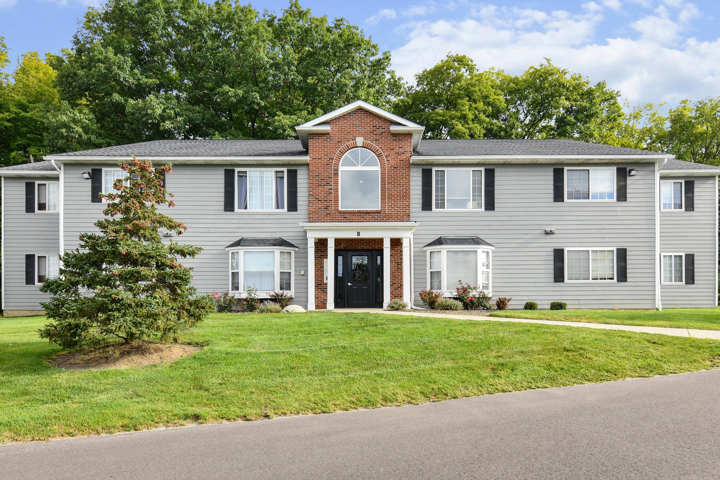 A two-story gray residential building with a brick accent and large front windows, surrounded by green grass and landscaping. A pathway leads to the entrance, which features a decorative door and window above. Trees provide a natural backdrop in the background under a clear blue sky.