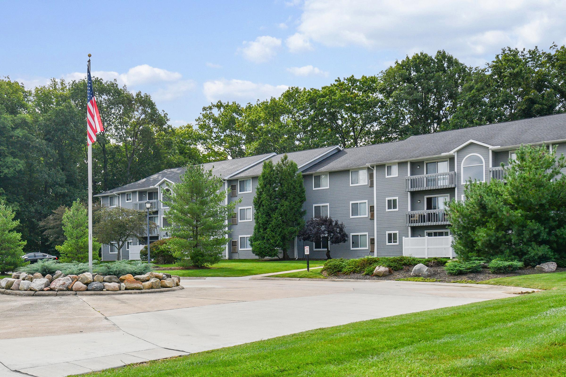 A modern apartment complex with gray siding and balconies surrounded by lush greenery. A circular stone garden in the foreground with a flagpole displaying the American flag. The sky is partly cloudy, and the environment appears well-maintained and inviting.