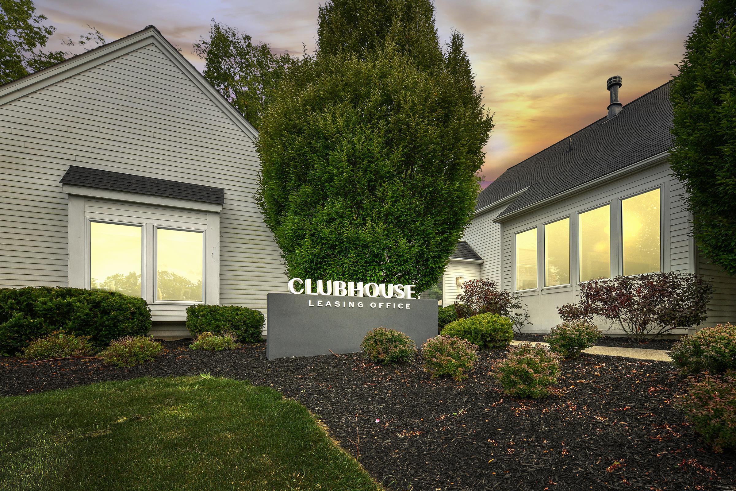 A leasing office building with a sign reading "CLUBHOUSE" is situated among well-maintained landscaping. The exterior features a combination of smooth grey siding and large windows, and the scene is illuminated by the warm glow of sunset. Lush green bushes and dark mulch enhance the aesthetic appeal.