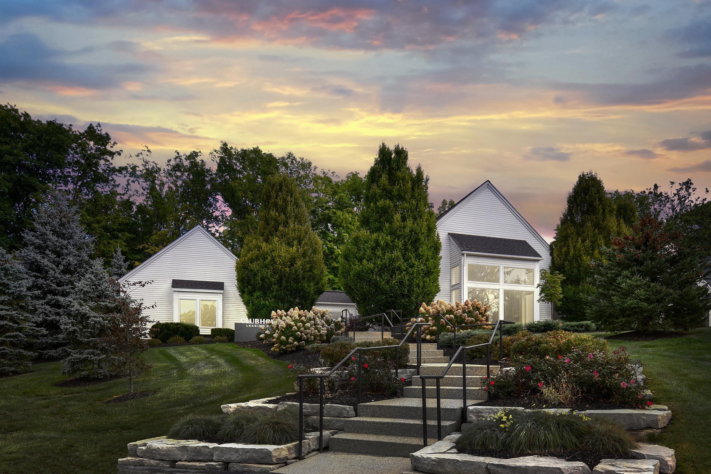 A serene landscape featuring two modern homes surrounded by lush greenery and colorful flowering plants. Stone steps lead up to the houses, complemented by a mix of trees and shrubs under a vibrant sunset sky. The scene conveys a sense of tranquility and inviting living spaces.