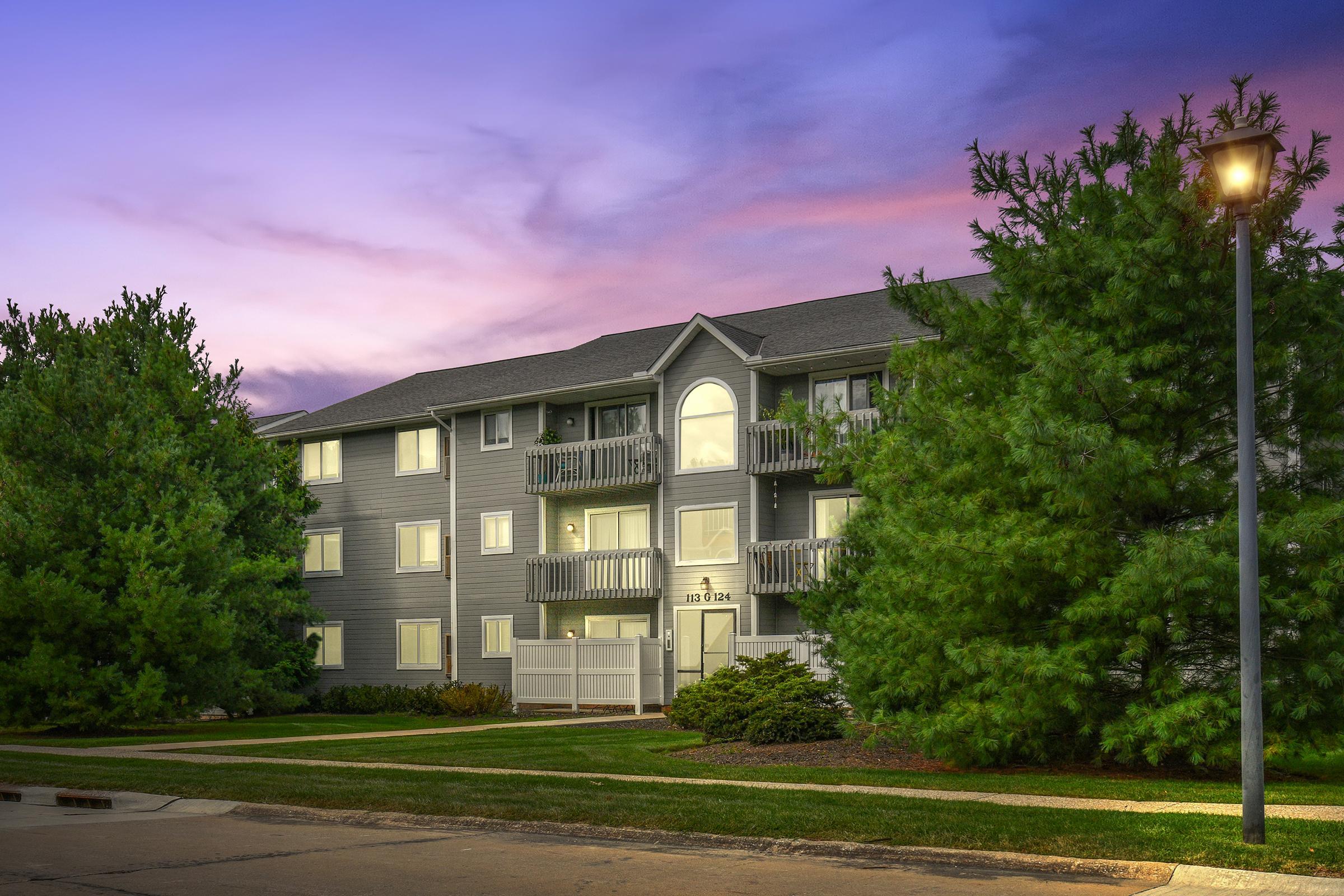 A modern apartment building with multiple balconies, surrounded by lush green trees, under a colorful sunset sky. A pathway leads to the entrance, and a lamp post is visible in the foreground, adding to the welcoming atmosphere.