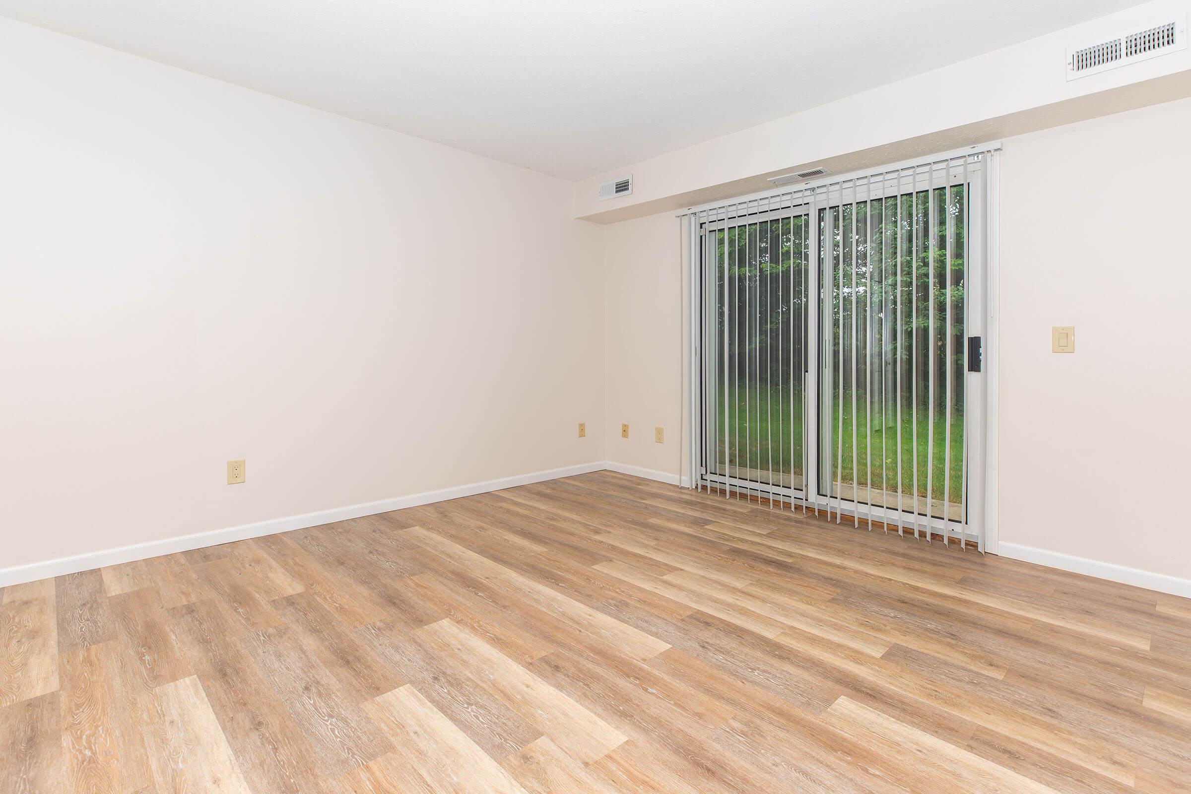 A spacious, empty room with light-colored walls and new wooden flooring. A large sliding glass door with vertical blinds leads to an outdoor area, allowing natural light to enter. The room appears clean and ready for furnishings, ideal for various design possibilities.