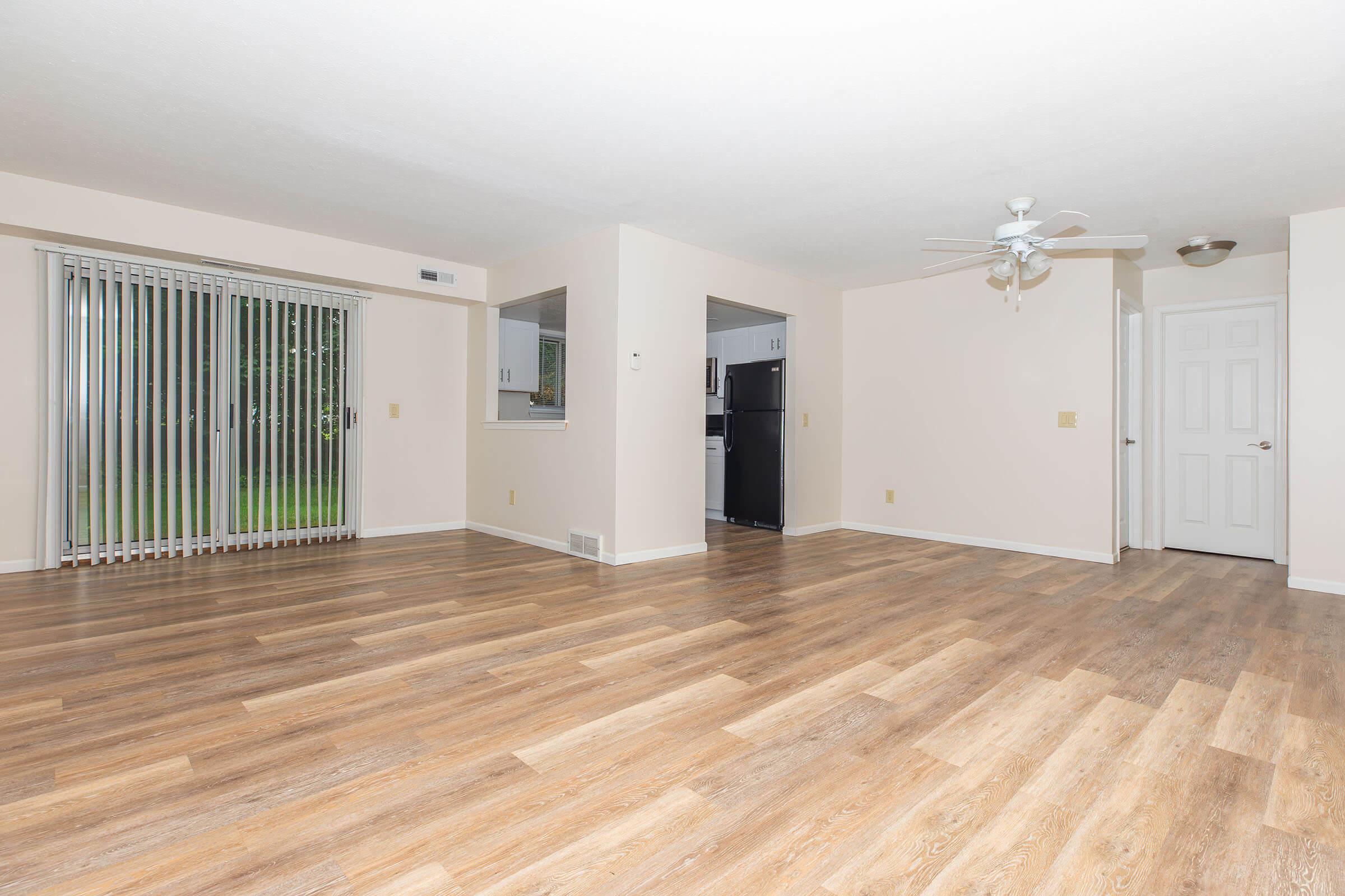 Spacious, light-filled living area with wood laminate flooring and sliding glass door leading to an outdoor space. A ceiling fan is installed, and there is an open view towards a kitchen area with appliances visible in the background. The walls are painted in a neutral color, creating a welcoming atmosphere.