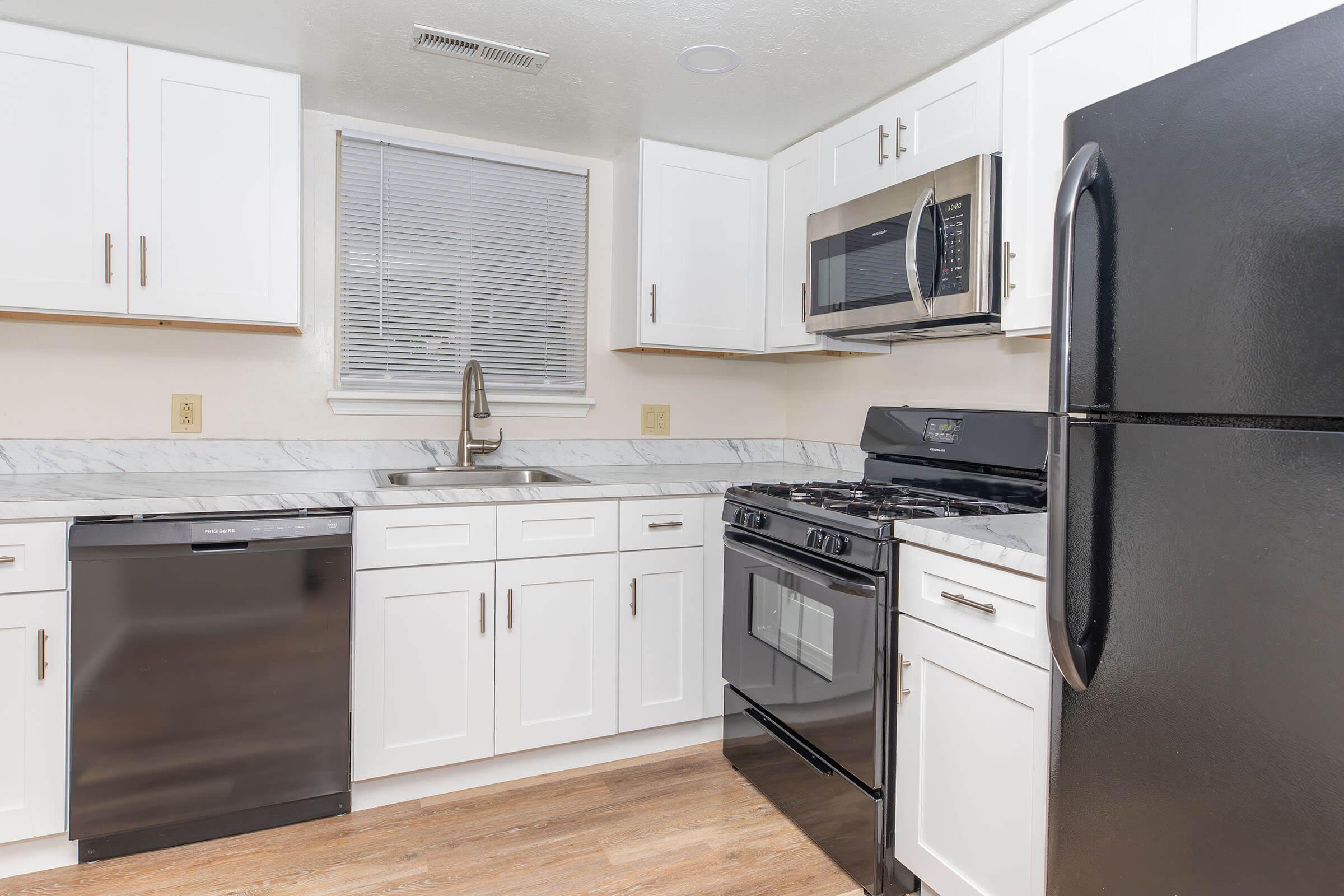 Modern kitchen featuring white cabinets, a stainless steel sink, and appliances including a microwave, gas stove, refrigerator, and dishwasher. The countertop is made of marble, and there's a window above the sink allowing natural light into the space. The flooring is a warm wood tone.