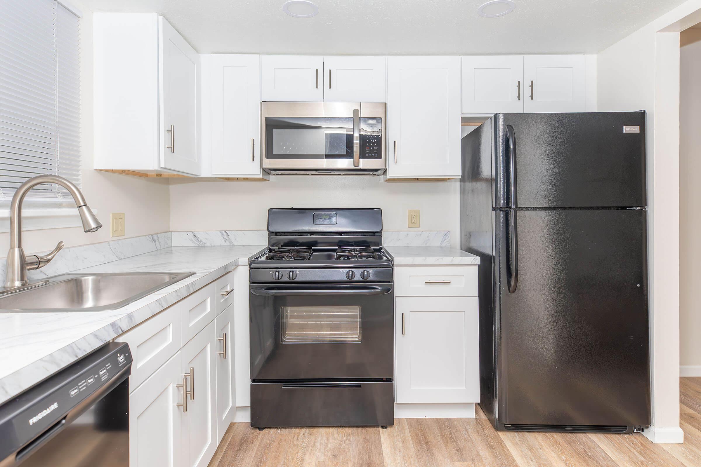 Modern kitchen featuring white cabinets, a stainless steel microwave and oven, a black gas stove, and a black refrigerator. The countertops are white marble, and the kitchen has a sink with a stylish faucet. The flooring is light wood.