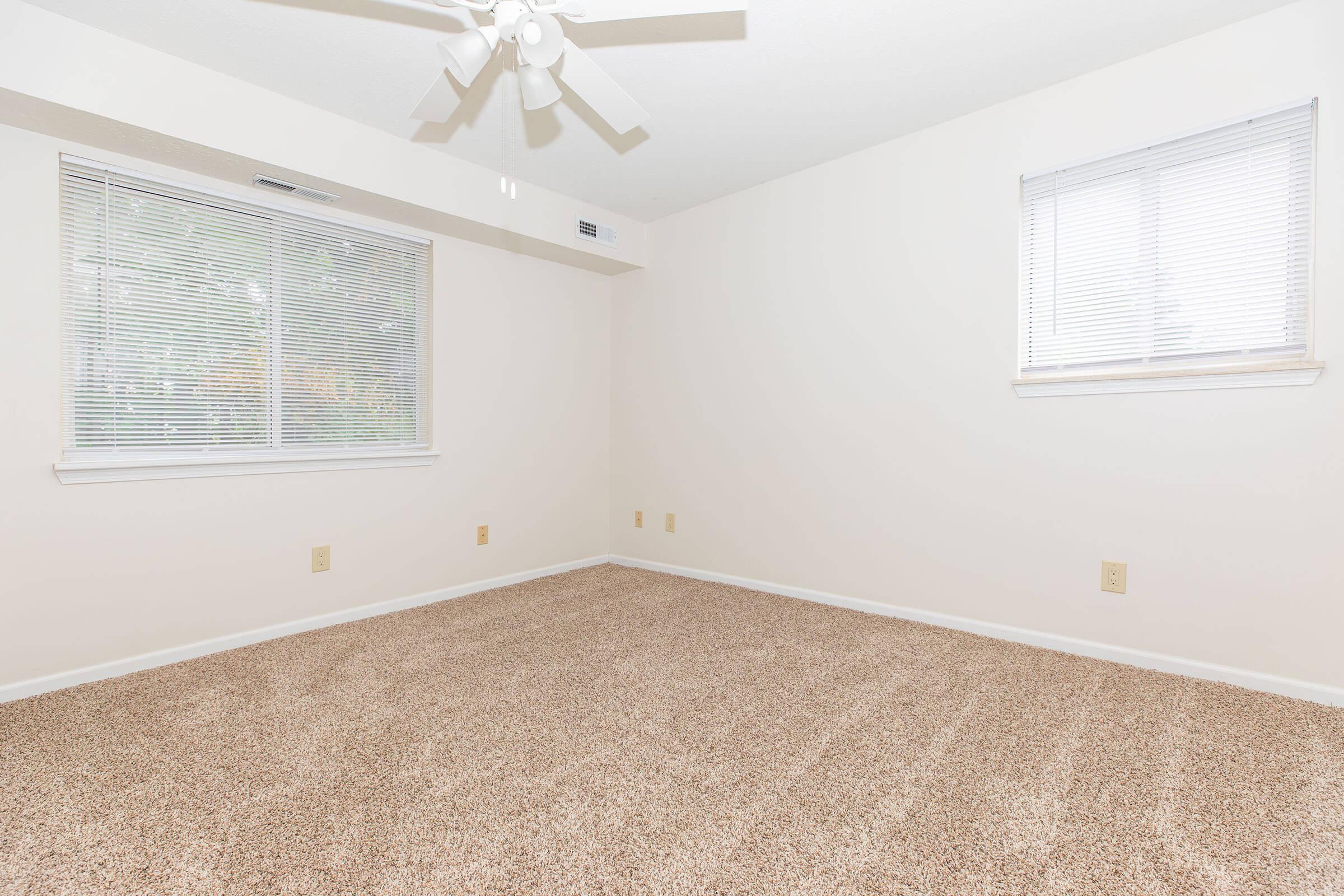 Empty room with beige carpet, light-colored walls, and a ceiling fan. The space has two windows with blinds, allowing natural light to enter, creating a bright and airy atmosphere. There are no furnishings or decorations present, giving the room a spacious and minimalist feel.